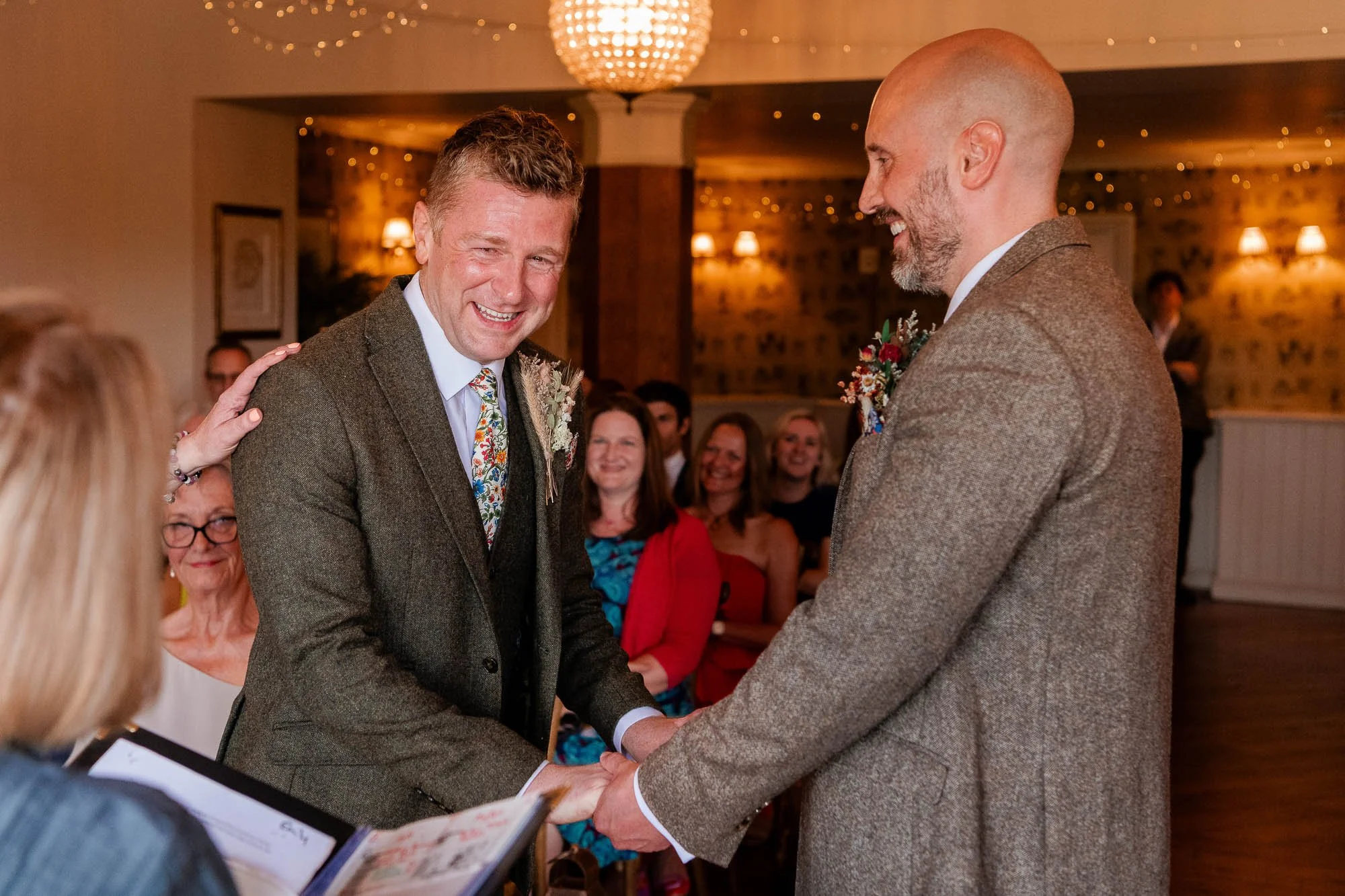 Two men in wedding suits holding hands during a wedding ceremony, smiling and facing each other, with friends and family in the background.