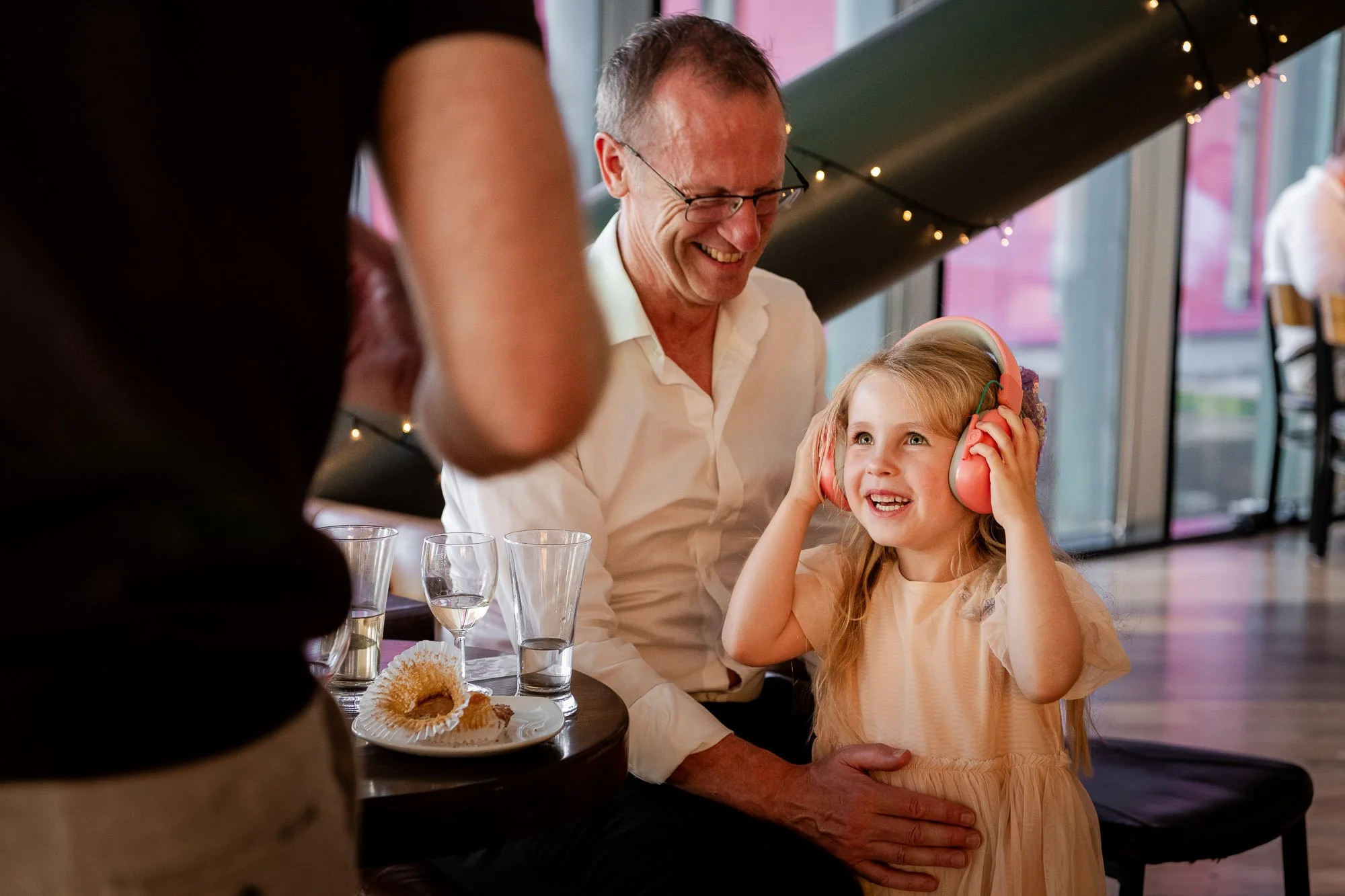 A joyful older man and young girl sitting at a table, the girl wearing pink headphones and smiling, with the man smiling alongside her. The table has glasses and a plate with food, and the scene is indoors with large windows and string lights in the 