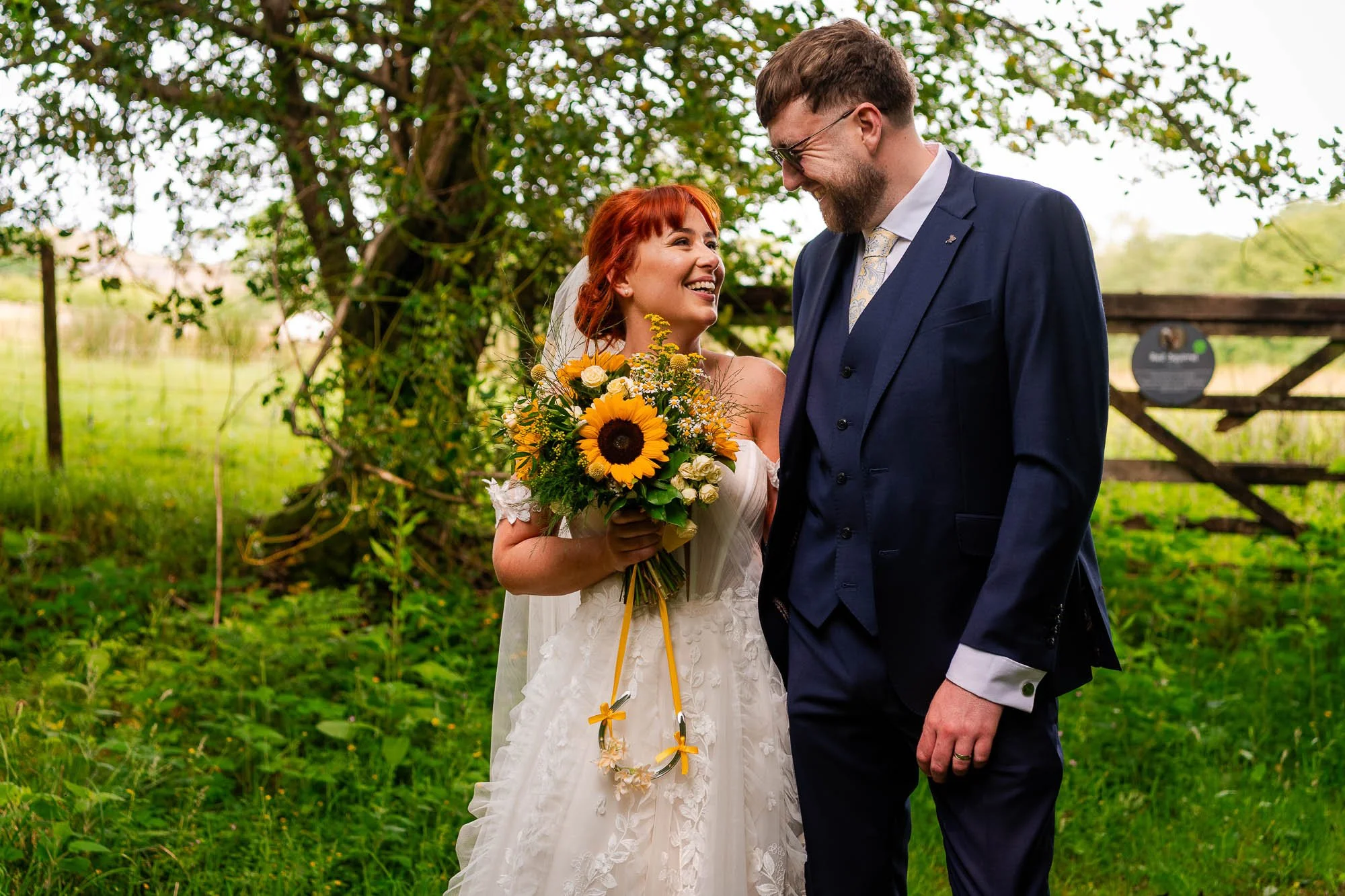 A bride in a white wedding dress holding a sunflower bouquet, smiling at a groom in a navy blue suit, outdoors near a tree and greenery during daytime.