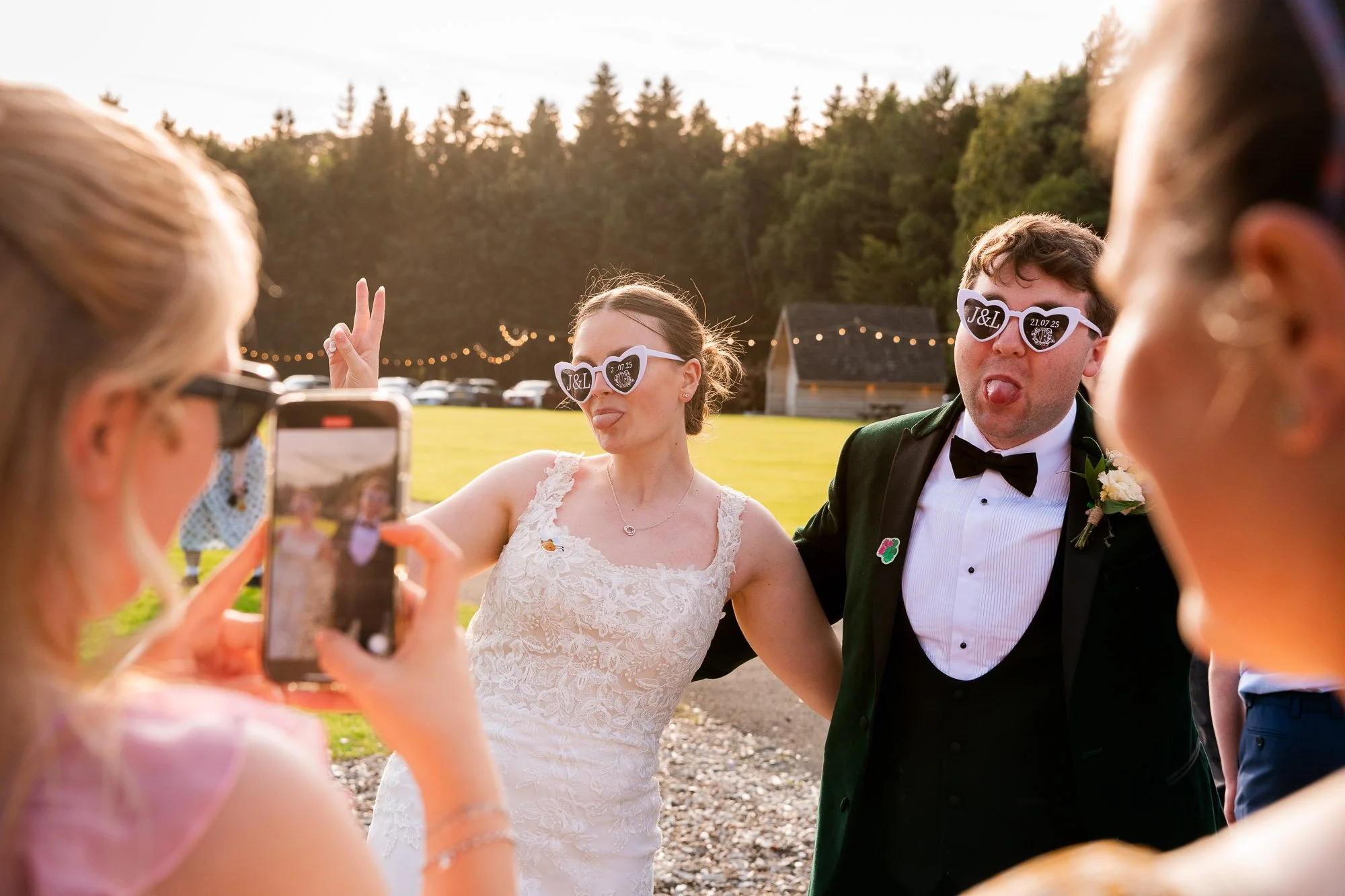 A bride and groom wearing sunglasses with 'J&L' and a date, posing playfully for photos at sunset during their outdoor wedding reception at Hobbit Hill, Clitheroe.