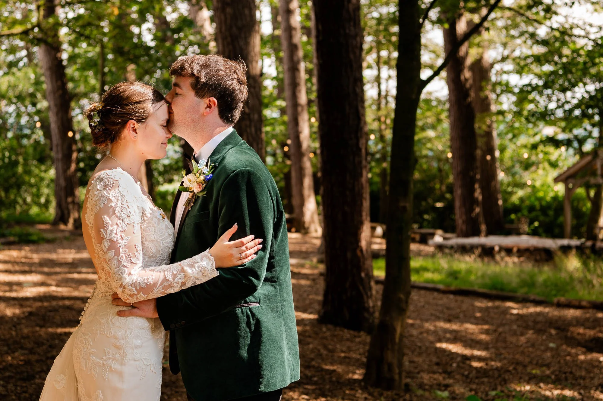 A bride and groom share a kiss in the forested ceremony space of Hobbit Hill, Clitheroe, with trees and sunlight in the background.