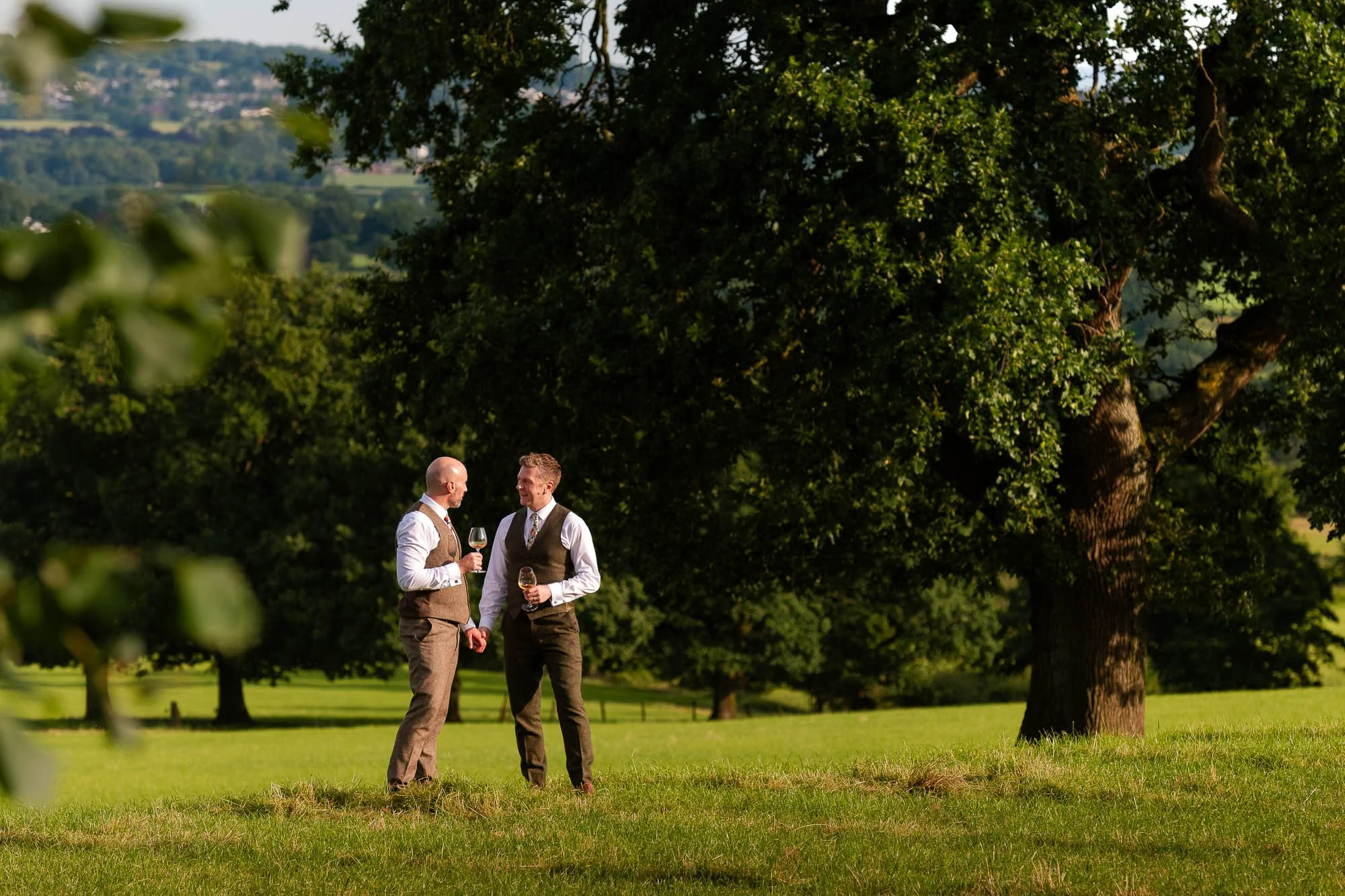 Two men wearing suits and ties, holding wine glasses, standing and talking outdoors on a grassy field with trees and a hilly landscape in the background.