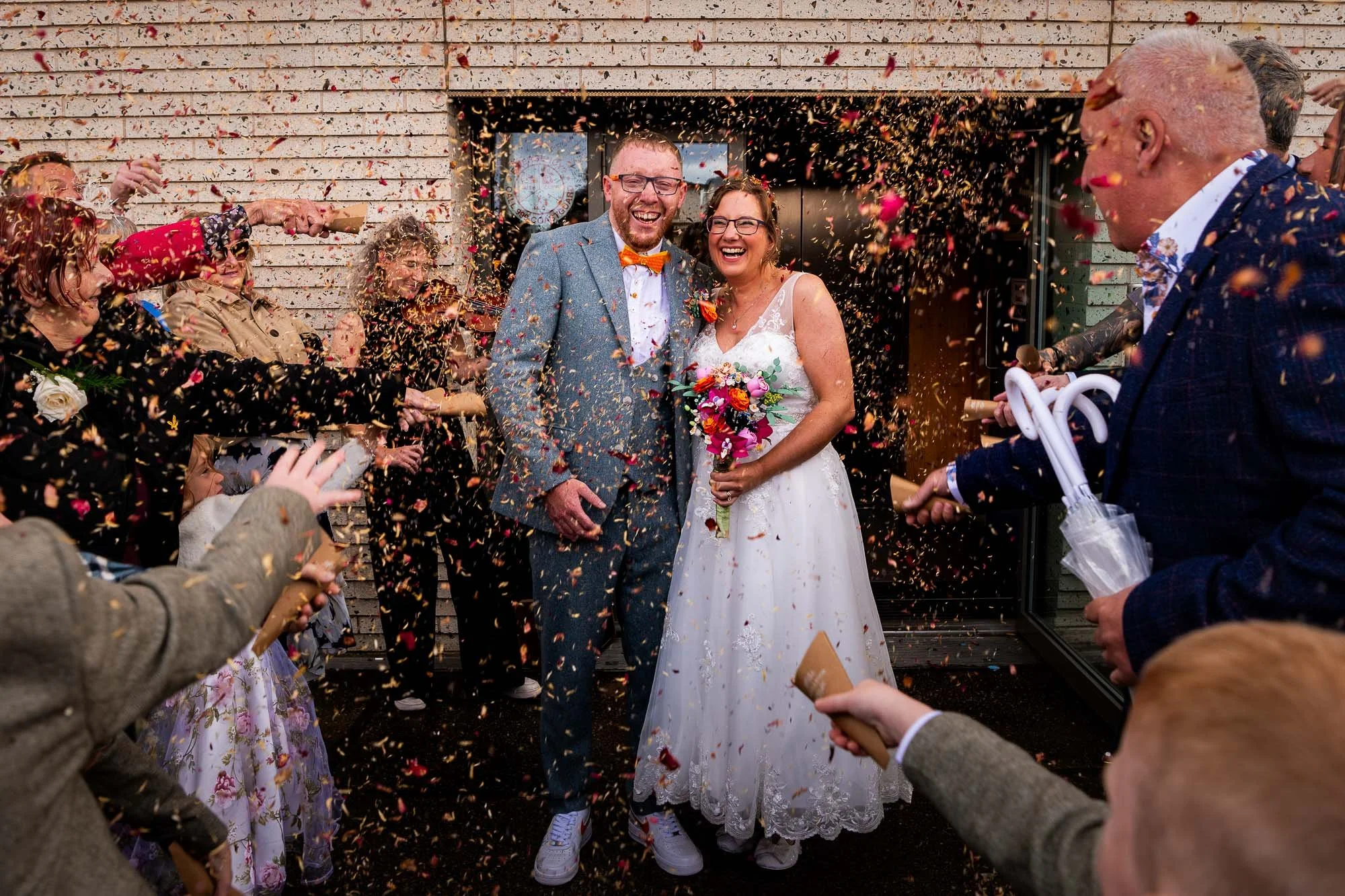 Happy newlywed couple celebrating outside as guests throw confetti, with bride holding a bouquet and wearing a white lace wedding dress, groom in a blue suit with a yellow bow tie, and people smiling.