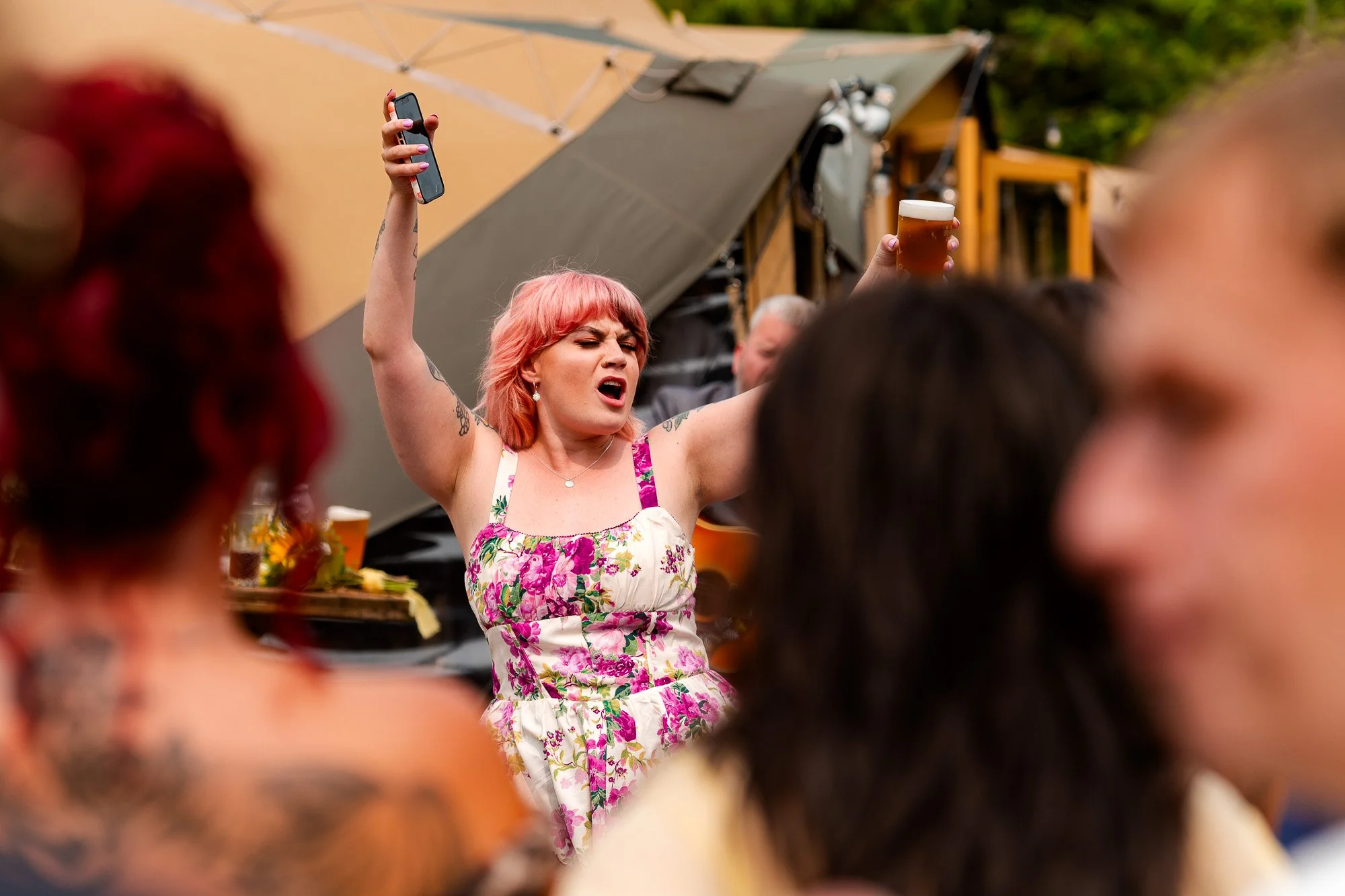 A woman with pink hair wearing a floral dress is singing passionately with her eyes closed and holding a beer. She is raising one arm and holding a phone in the other hand at an outdoor gathering or concert.