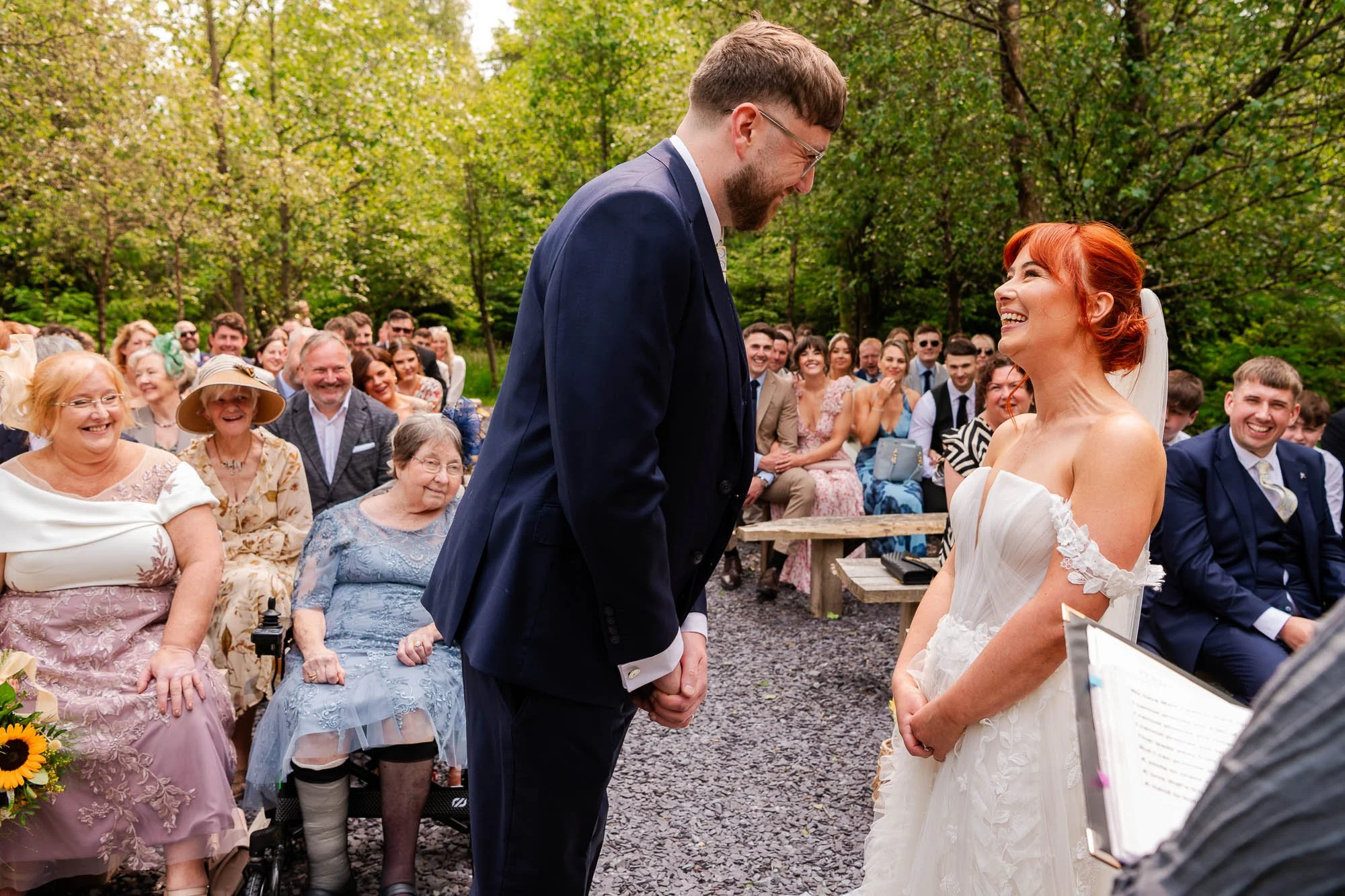 A bride and groom exchanging vows during an outdoor wedding ceremony, with the groom bowing his head and holding the bride's hands, while the bride smiles at him. Guests seated behind them are smiling and watching, surrounded by greenery.
