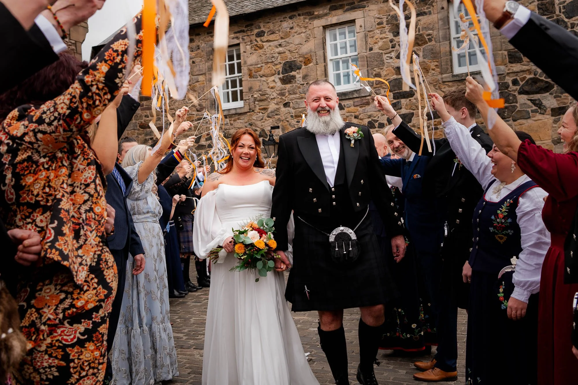 Bride and groom walking under a confetti arch, surrounded by wedding guests in traditional Scottish dress, outdoors with stone building in background.