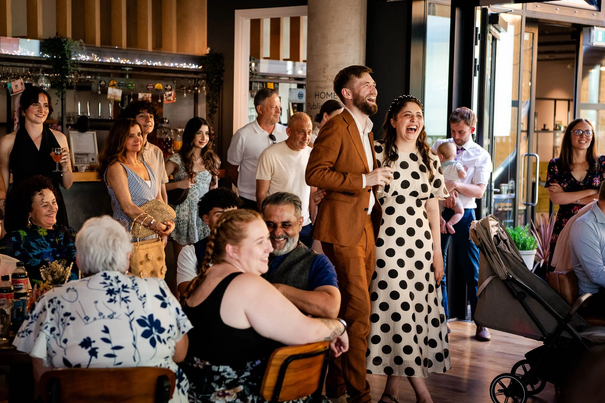 People smiling and laughing at an indoor social gathering or celebration, with a mix of seated and standing individuals.