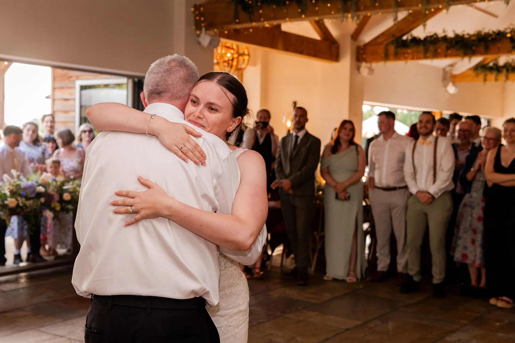 A bride and groom share a heartfelt dance with guests watching in a warmly lit hall decorated with flowers and greenery.