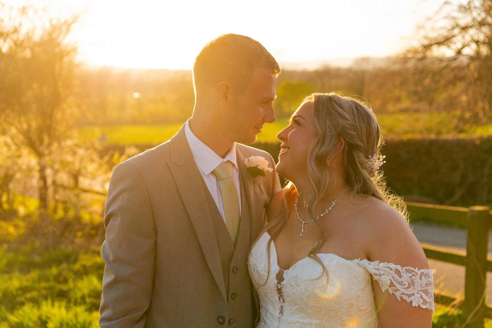 A newlywed couple facing each other outdoors at sunset, smiling with noses almost touching, in a scenic setting with trees and a wooden fence.