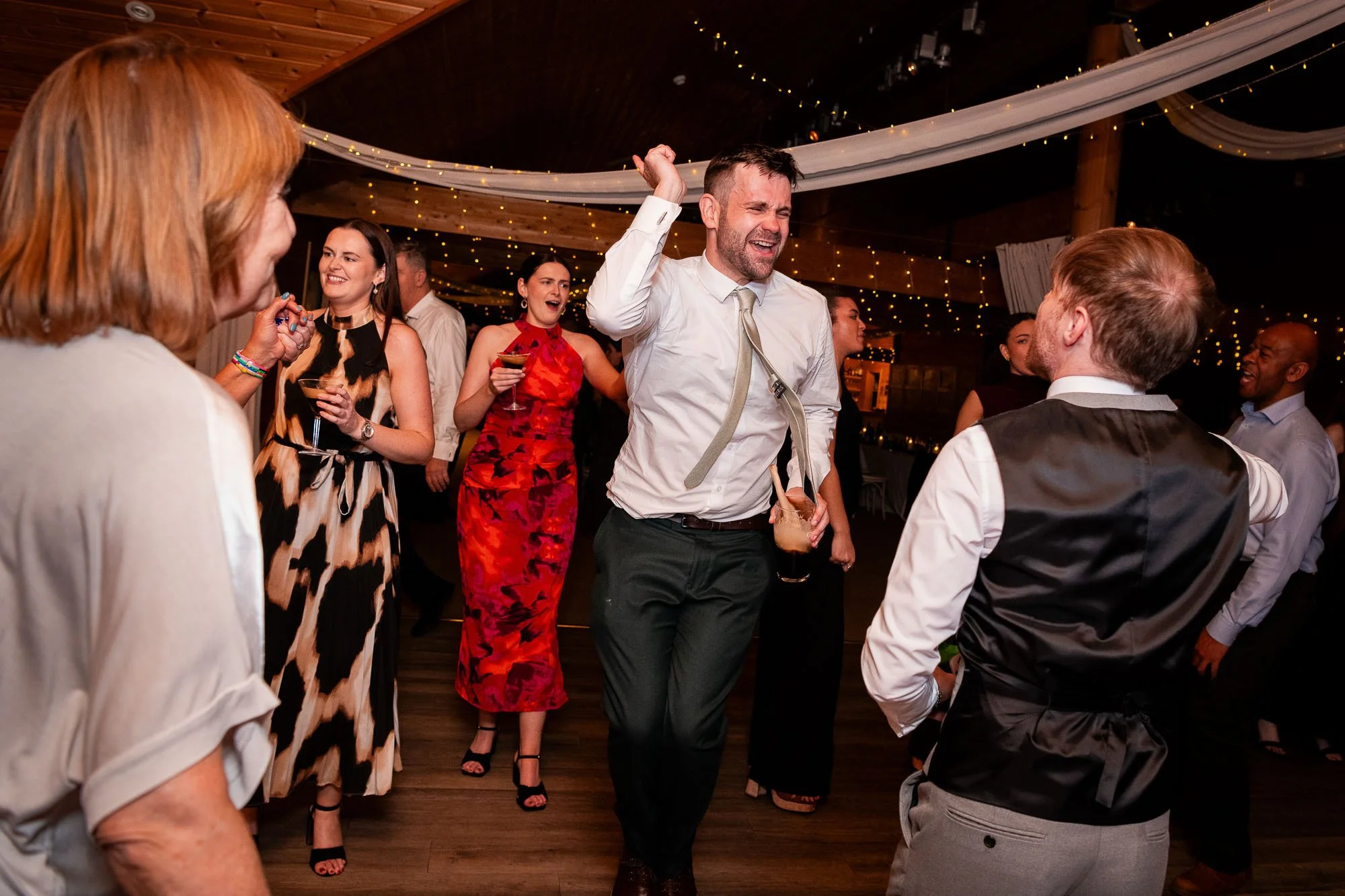 People dancing and celebrating at a party or wedding reception indoors, under string lights and draped fabric on the ceiling.