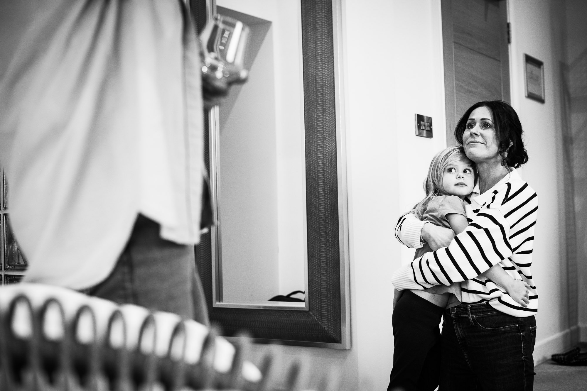 A woman is holding a young girl in an interior hallway, with a person partially visible in the foreground. The woman and girl are looking at the person in the foreground, with the woman wearing a striped shirt and the girl appearing to be about 4-6 y