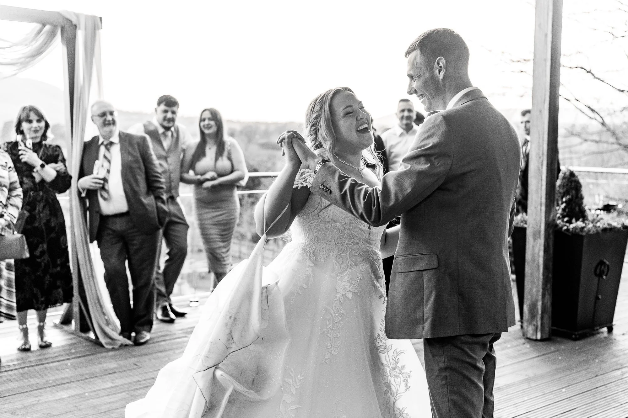 A bride and groom dancing at their wedding reception with friends and family watching, set outdoors on a wooden deck.