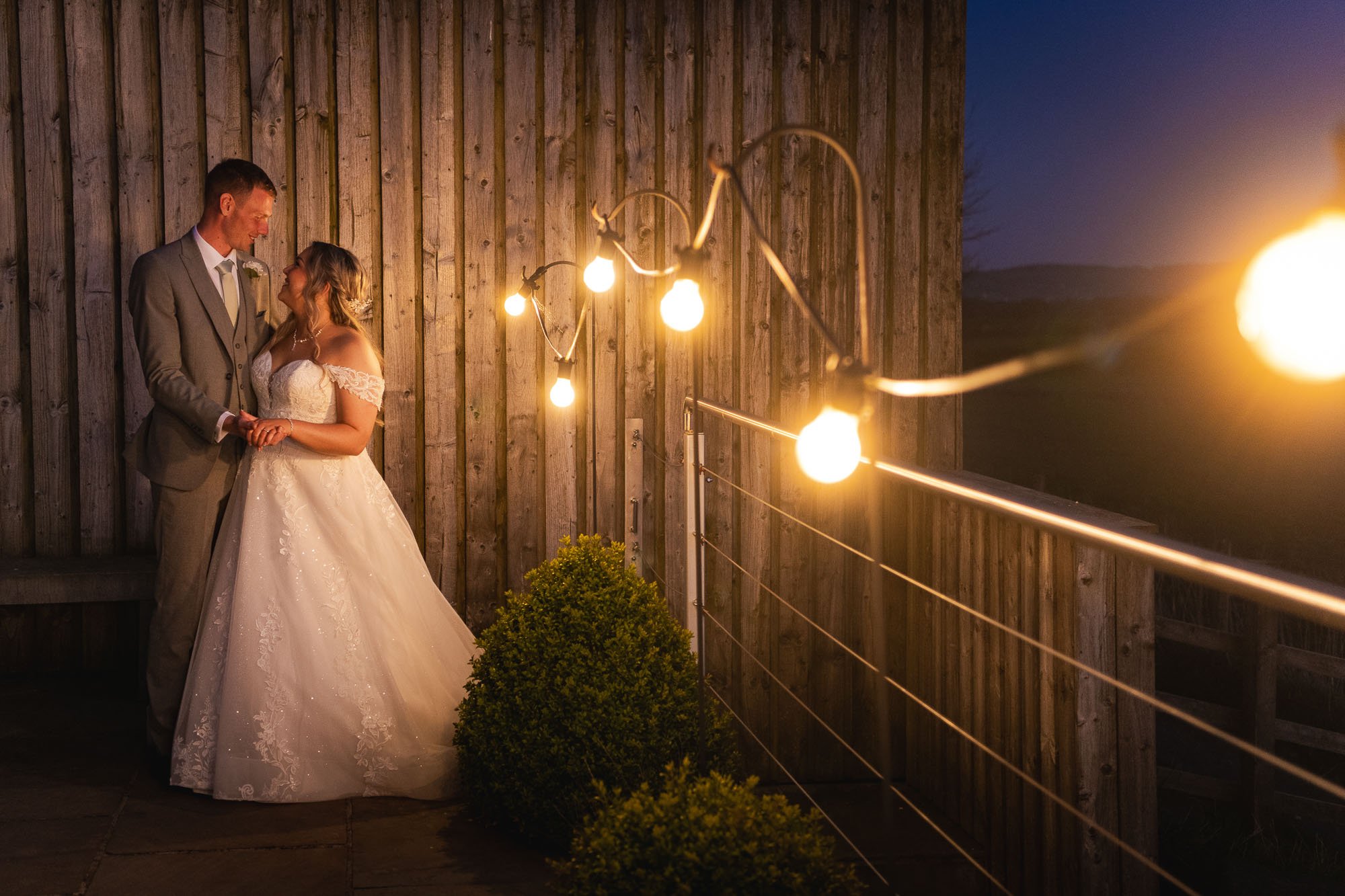 A bride and groom holding hands, standing close together, illuminated by string lights on a wooden outdoor deck during dusk.