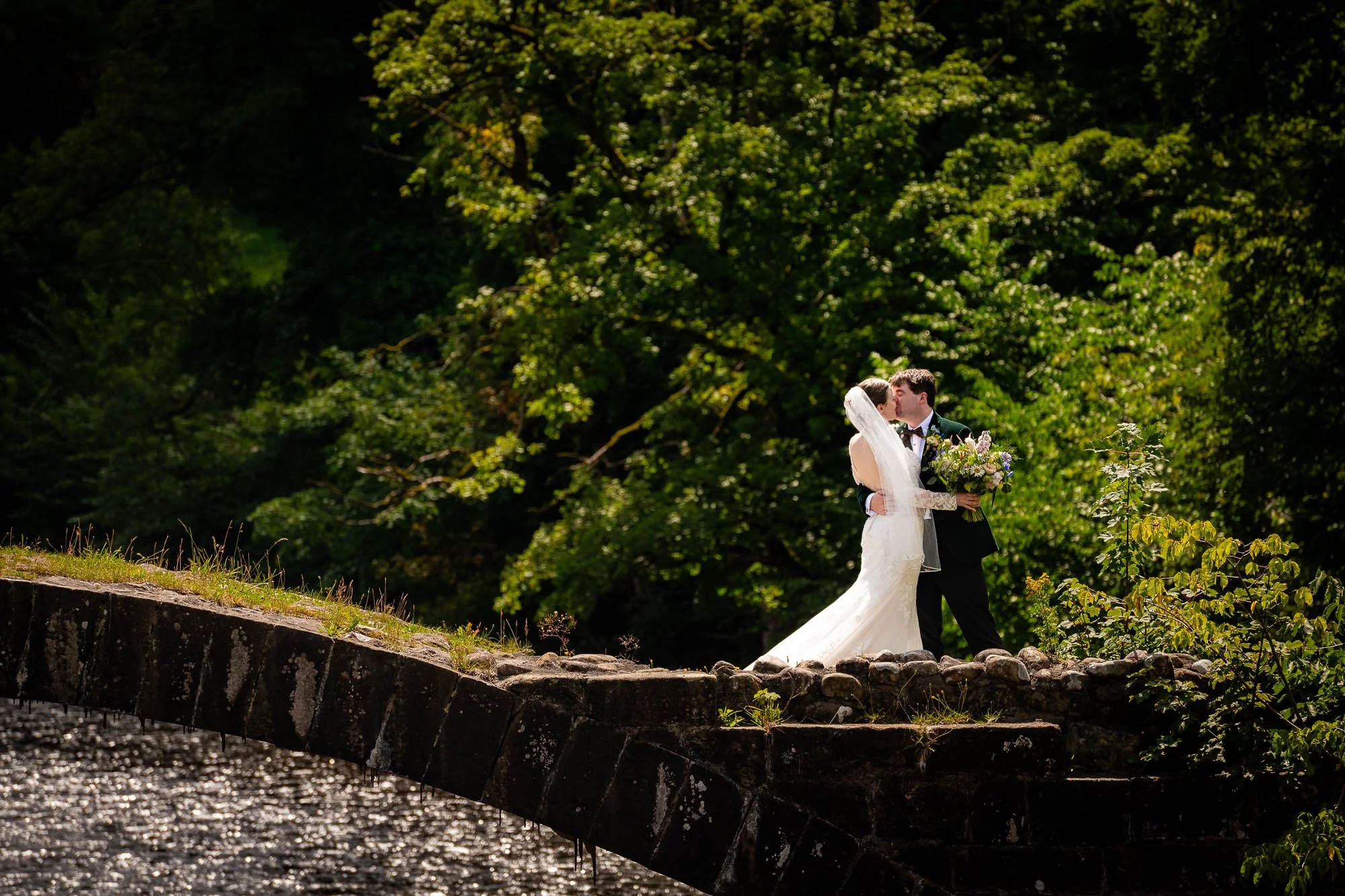 A bride and groom sharing a kiss outdoors during their wedding, with a lush green background and sunlight highlighting the scene in Lancashire.