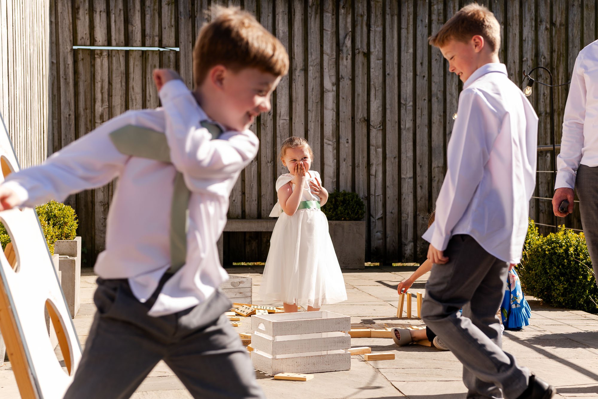 Children playing a game outdoors, with a young girl in a white dress covering her nose in the background and boys running in the foreground.