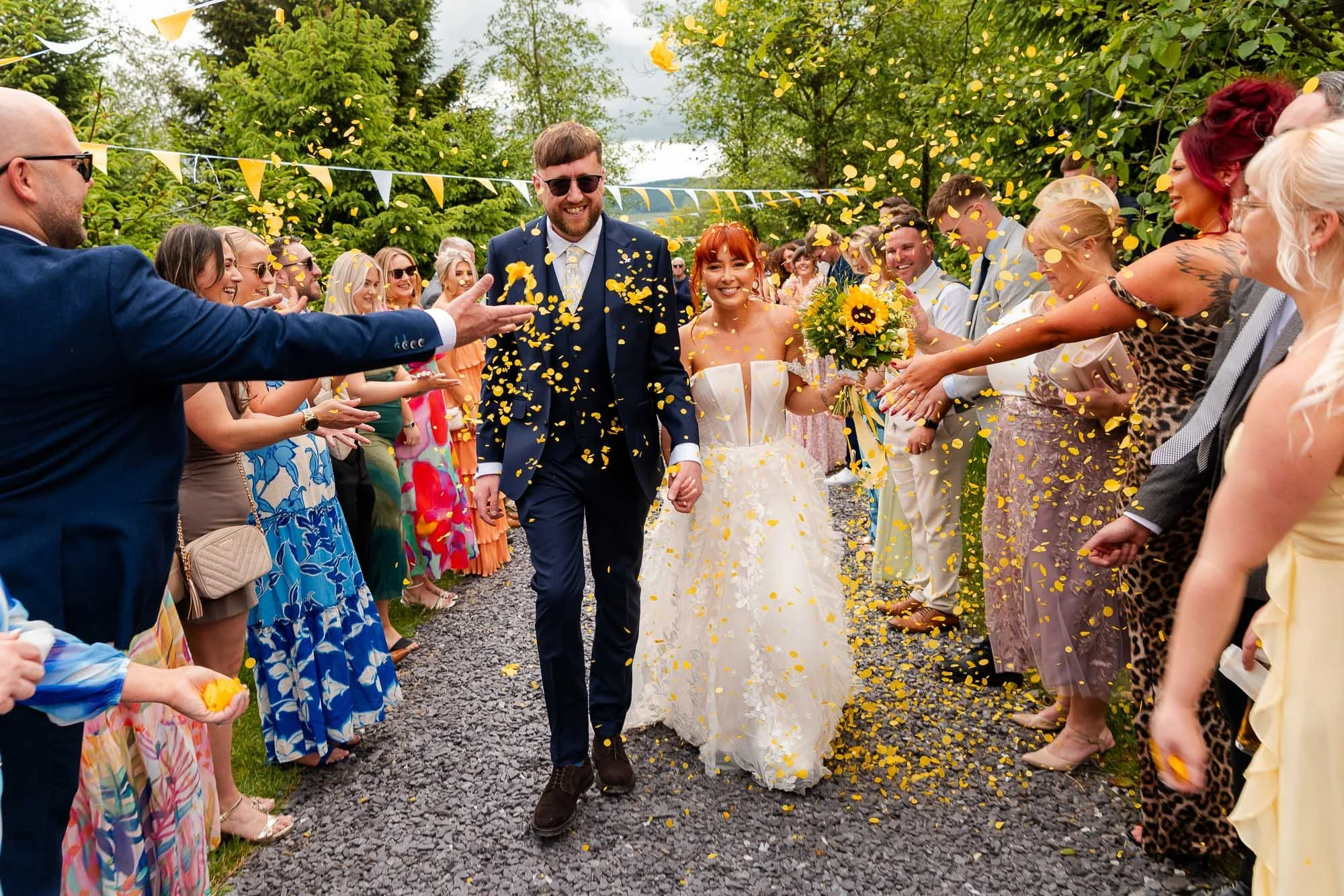 A newly married couple walking down a celebratory outdoor aisle lined with friends and family, confetti falling around them, during daytime.