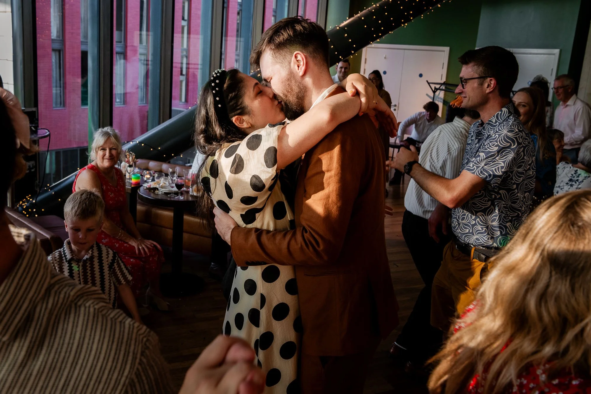 A couple is dancing and kissing at a celebration party with friends and family, with some sitting and some standing, inside a colorful venue with large windows and string lights.