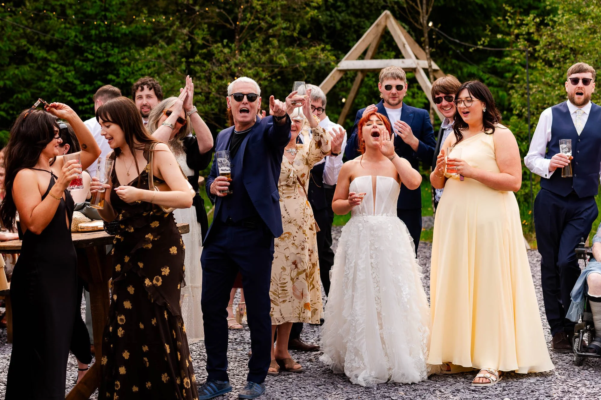 Group of people at an outdoor celebration, some dancing and holding drinks, with trees and a wooden structure in the background.