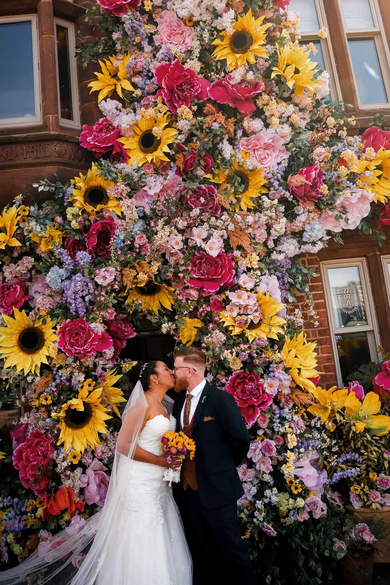 Bride and groom kissing in front of a wall of colorful flowers, including sunflowers, peonies, and roses, at a wedding celebration in Manchester.