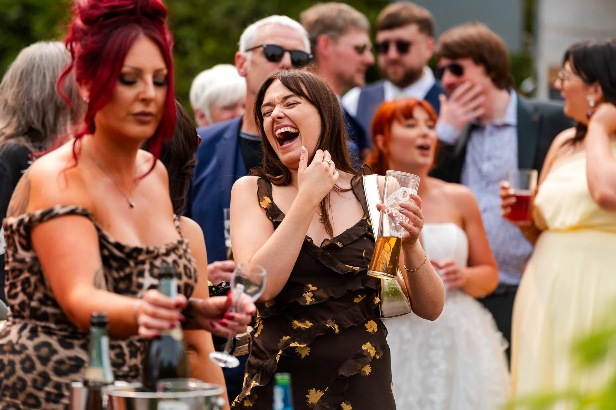 Group of people at an outdoor party or wedding reception, some laughing and holding drinks, with a woman in the center laughing and holding a pint glass.
