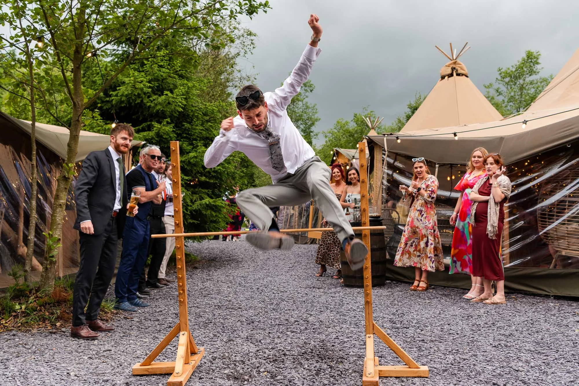 A man in a white shirt and gray pants is jumping over a slackline at an outdoor event, surrounded by onlookers dressed in casual and semi-formal attire under a cloudy sky.