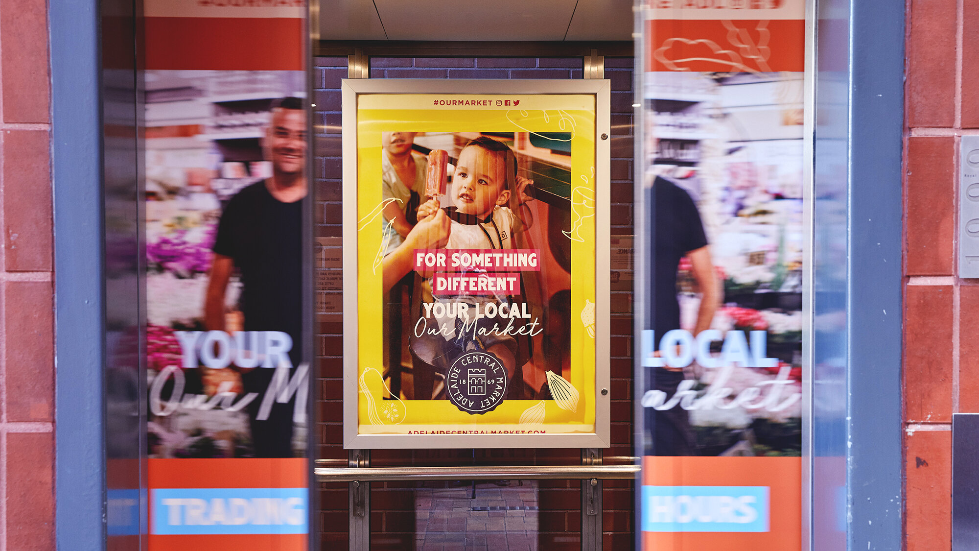 A colorful poster inside a subway station advertising a local market in Adelaide, featuring a young girl eating a popsicle. The poster has a yellow background with white and pink text that reads, 'For something different, your local Our Market,' along with the Adelaide Central Market logo and website. The subway doors reflect blurred images of people and the surrounding area.