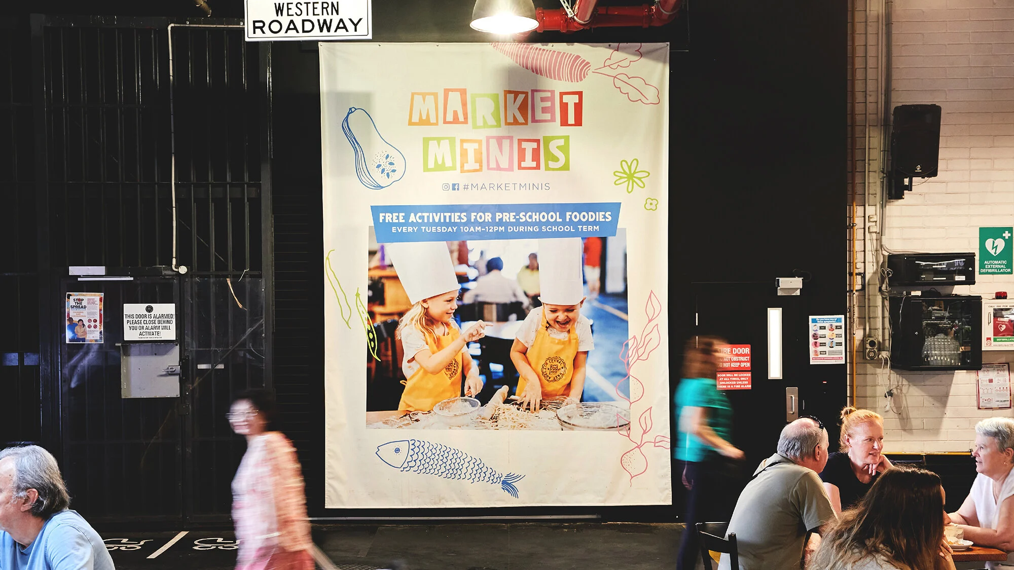 Large promotional banner for Market Minis event featuring children dressed as chefs cooking, with text about free activities for pre-school children every Tuesday from 10 AM to 12 PM, inside a busy indoor space with people dining.