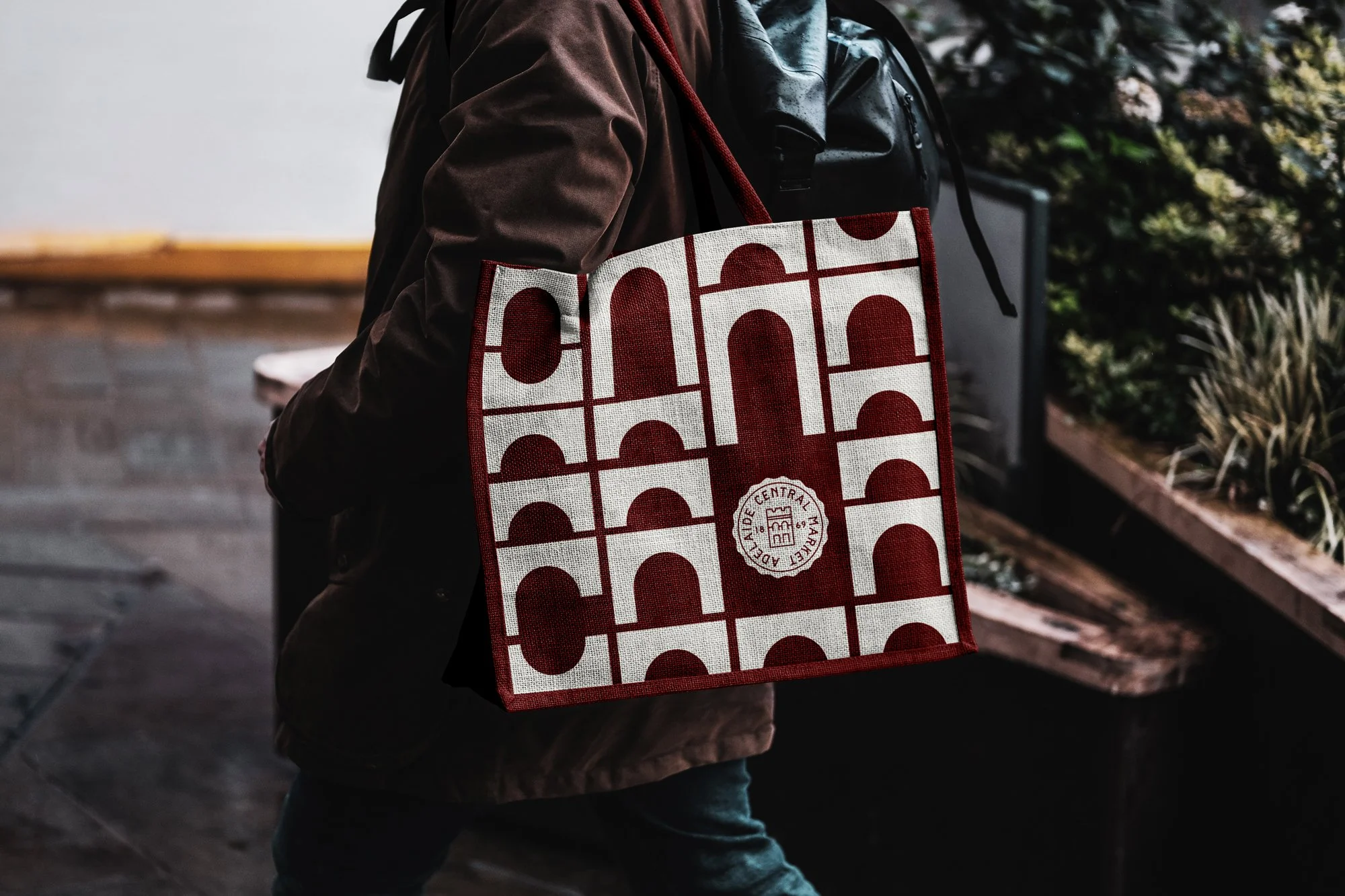 Person carrying a red and white patterned tote bag with a circular logo, walking indoors near a display of plants.