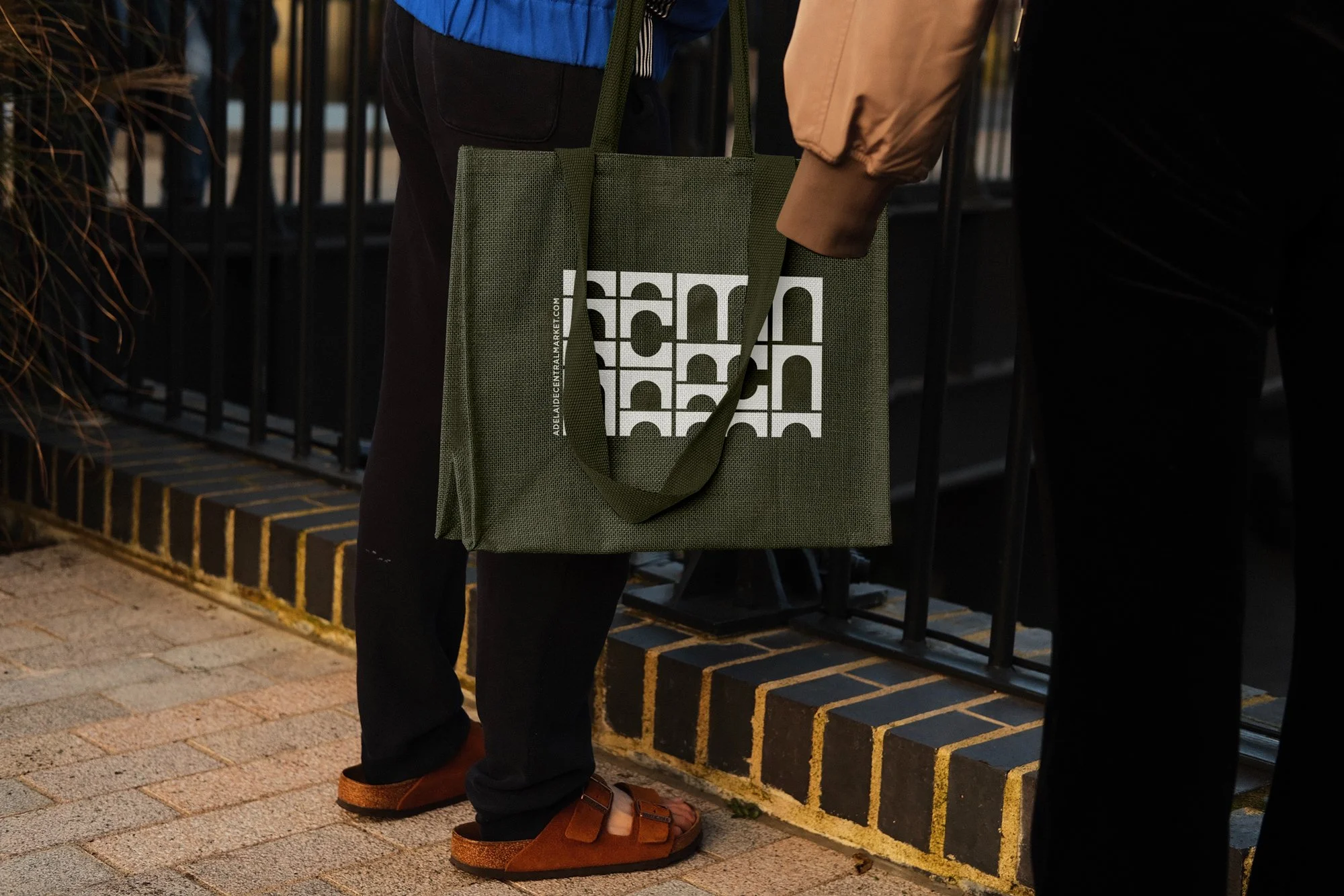 Person standing outdoors holding a green reusable shopping bag with black branding. The person is wearing brown sandals, black pants, and a tan jacket, with a blue jacket visible atop. The scene includes a brick sidewalk, a black metal fence, and another person partially visible to the right.