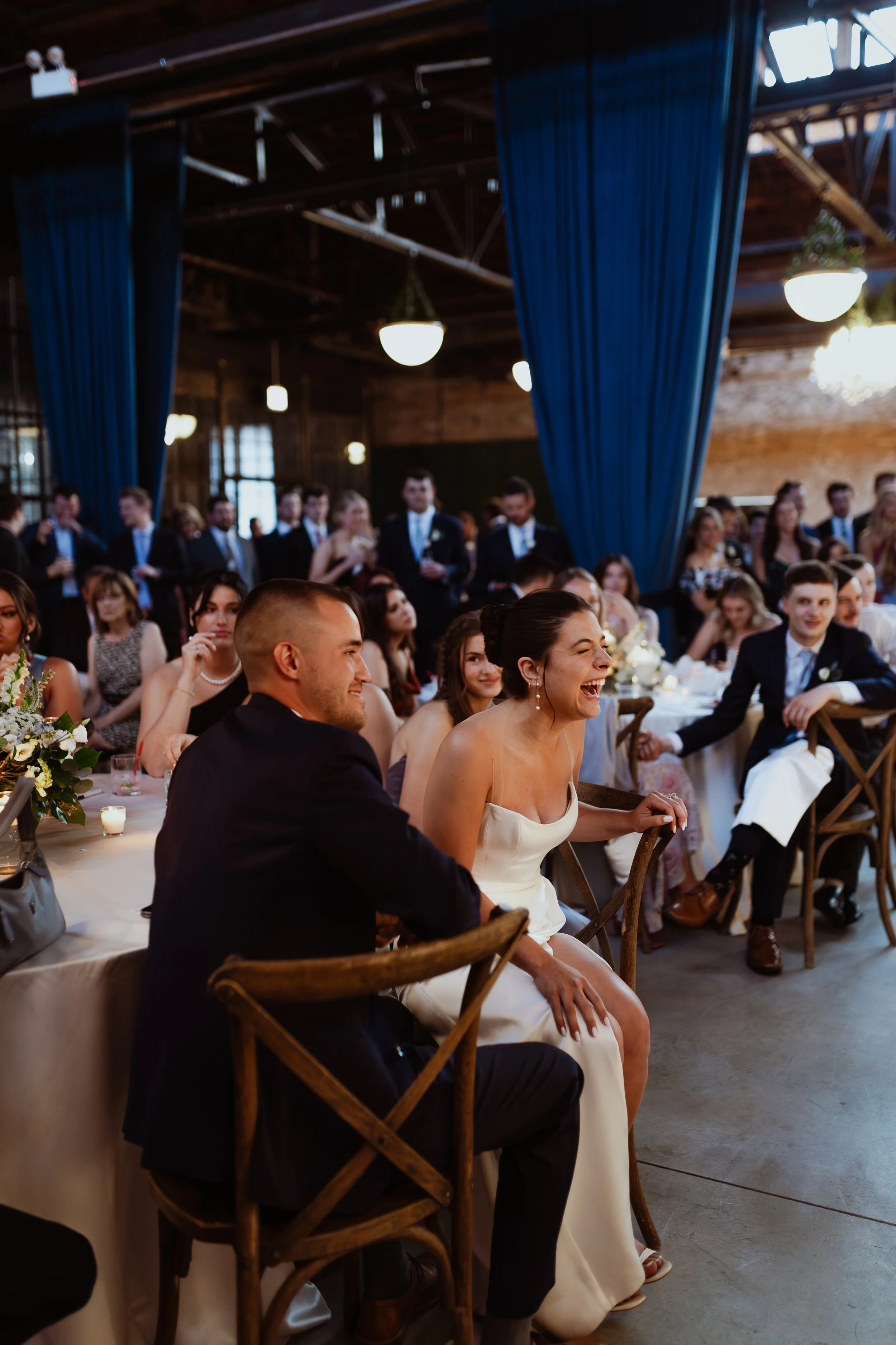 People at a wedding reception at Wildman BT Chicago, sitting at tables, with some standing and talking in the background, inside a rustic wooden venue with blue curtains and hanging lights. 