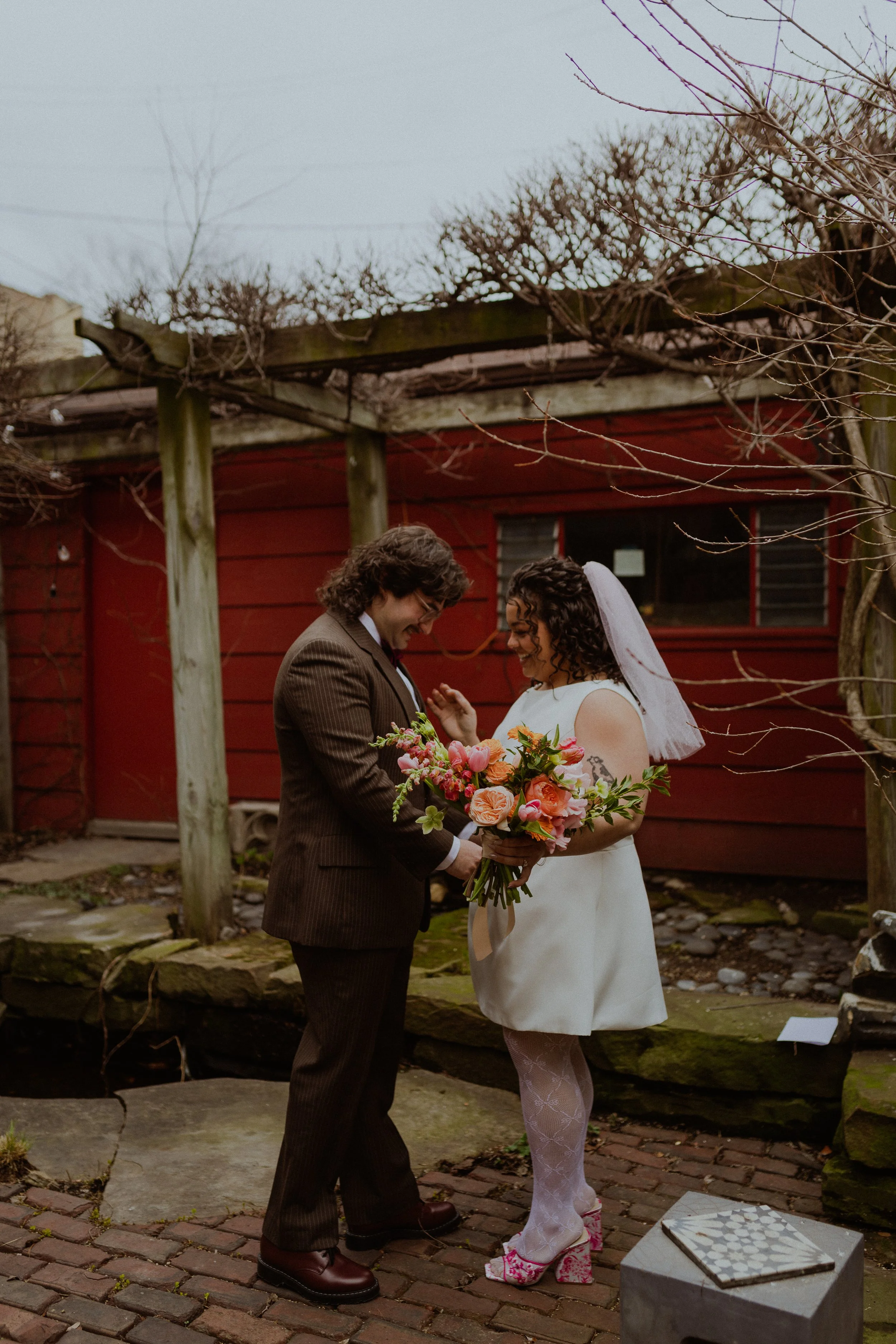 A couple dressed in wedding attire standing on a brick path outdoors, exchanging rings while holding a bouquet of pink and orange flowers, with a red building and bare trees in the background. Chicago Micro Wedding First Look Logan Square Airbnb