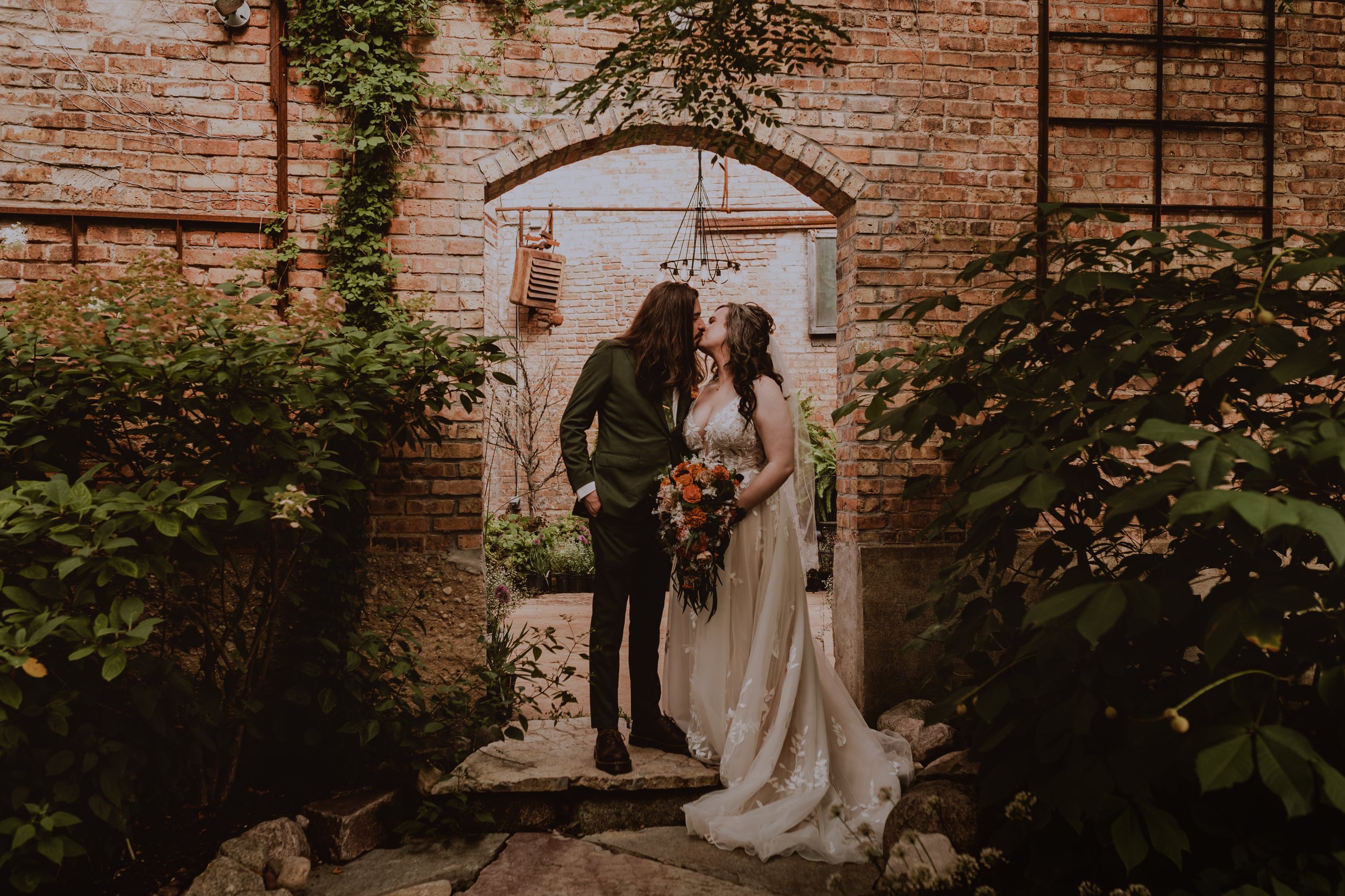 Two women in wedding attire sharing a kiss under an archway in a brick courtyard, with one woman holding a bouquet of flowers.