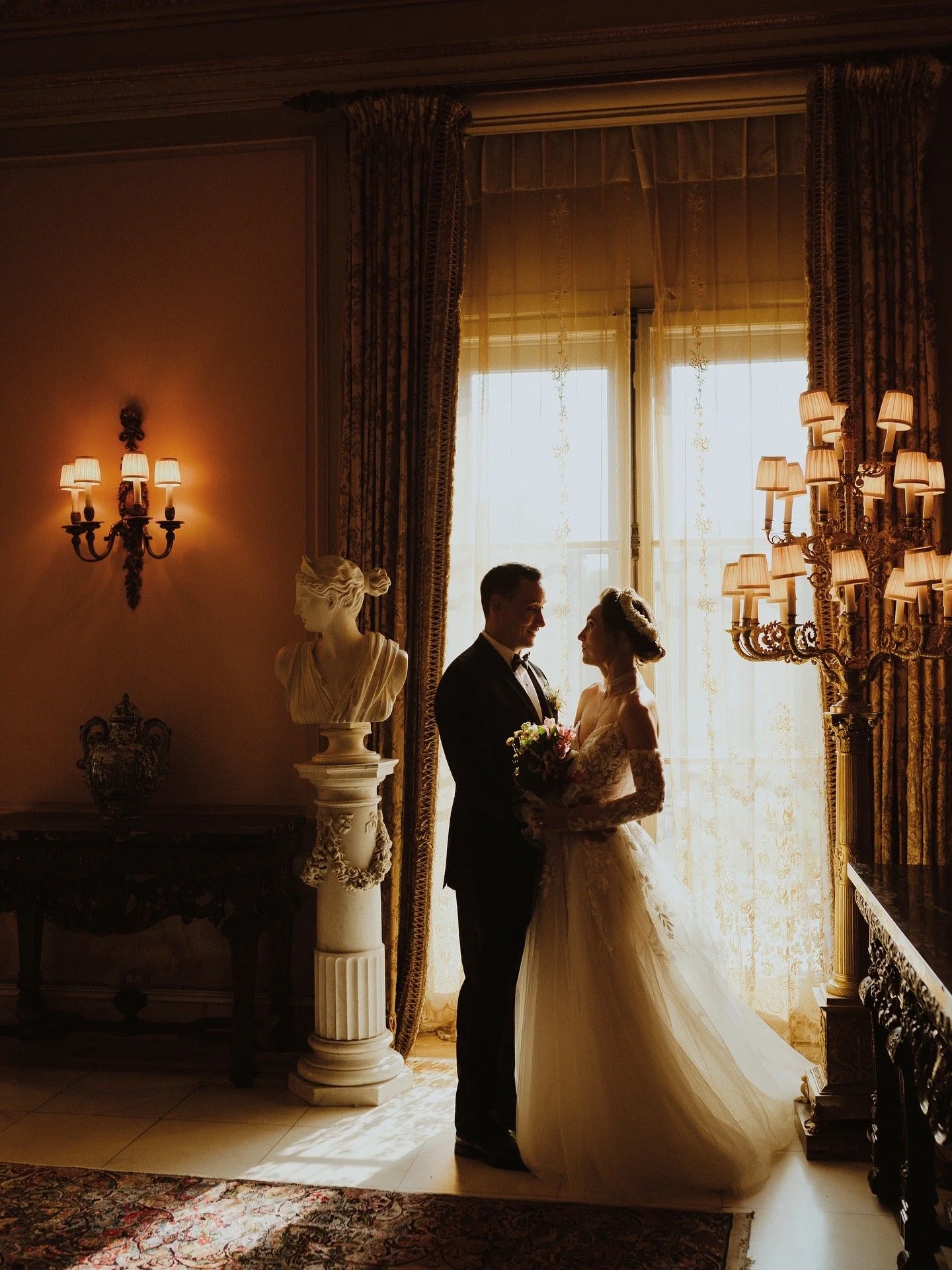 A bride and groom face each other in silhouette in a room with elegant curtains, ornate wall sconces, and classic decor, illuminated by soft natural light from large windows. Editorial Wedding Portraits Couple at Cuneo mansion.