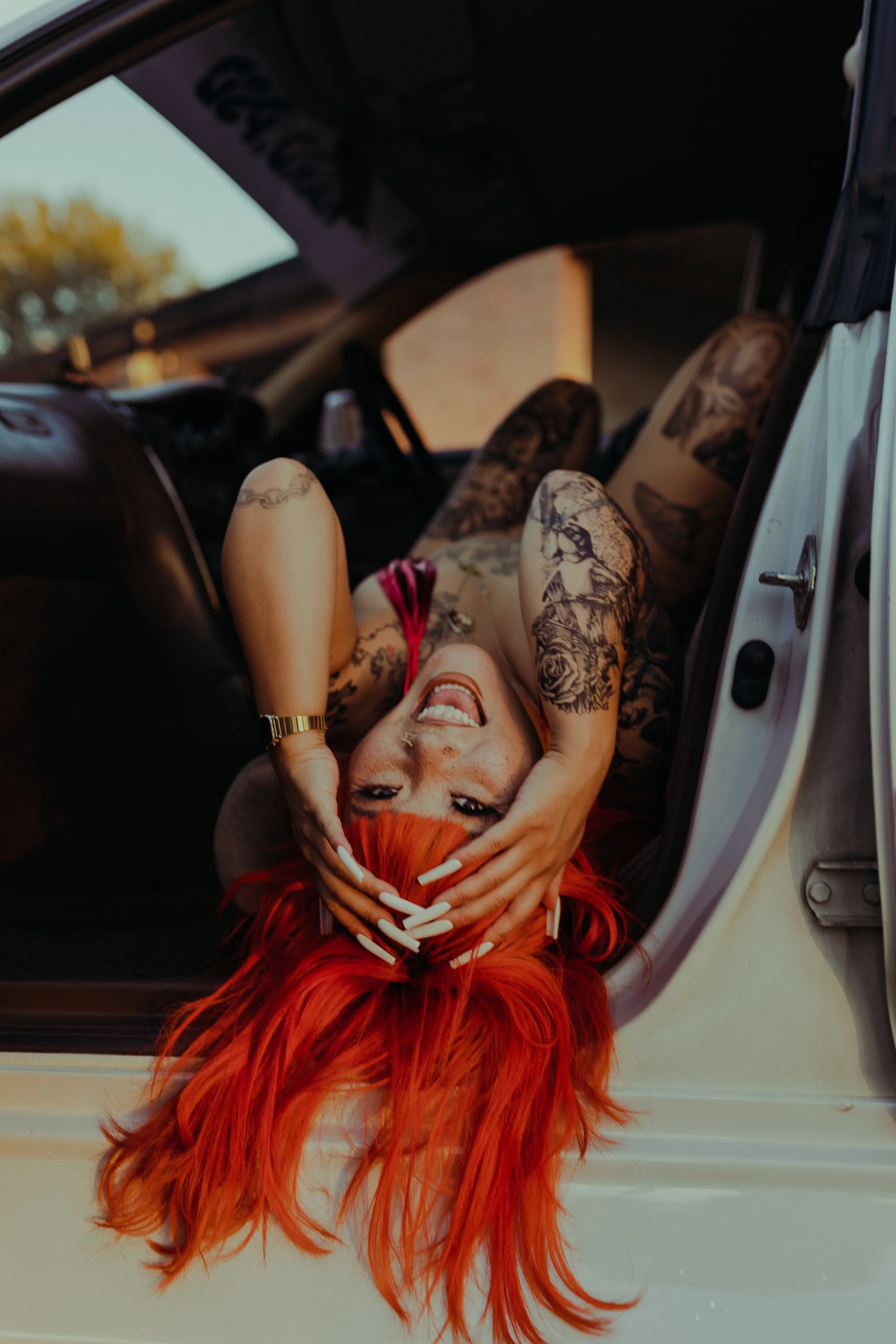 Model with red/orange hair laying upside down across the front seat of a white lowered drift car in a red one piece bathing suit covered in tattoos with long white square nails by Ameya
