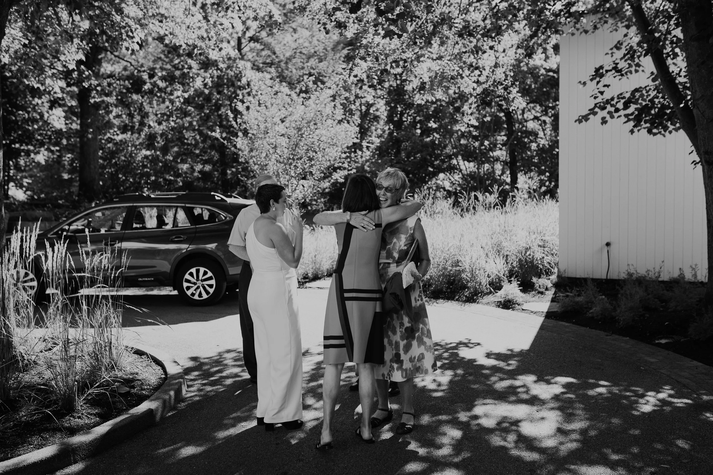 Four women hugging and talking outdoors on a sunny day, with a car and trees in the background.