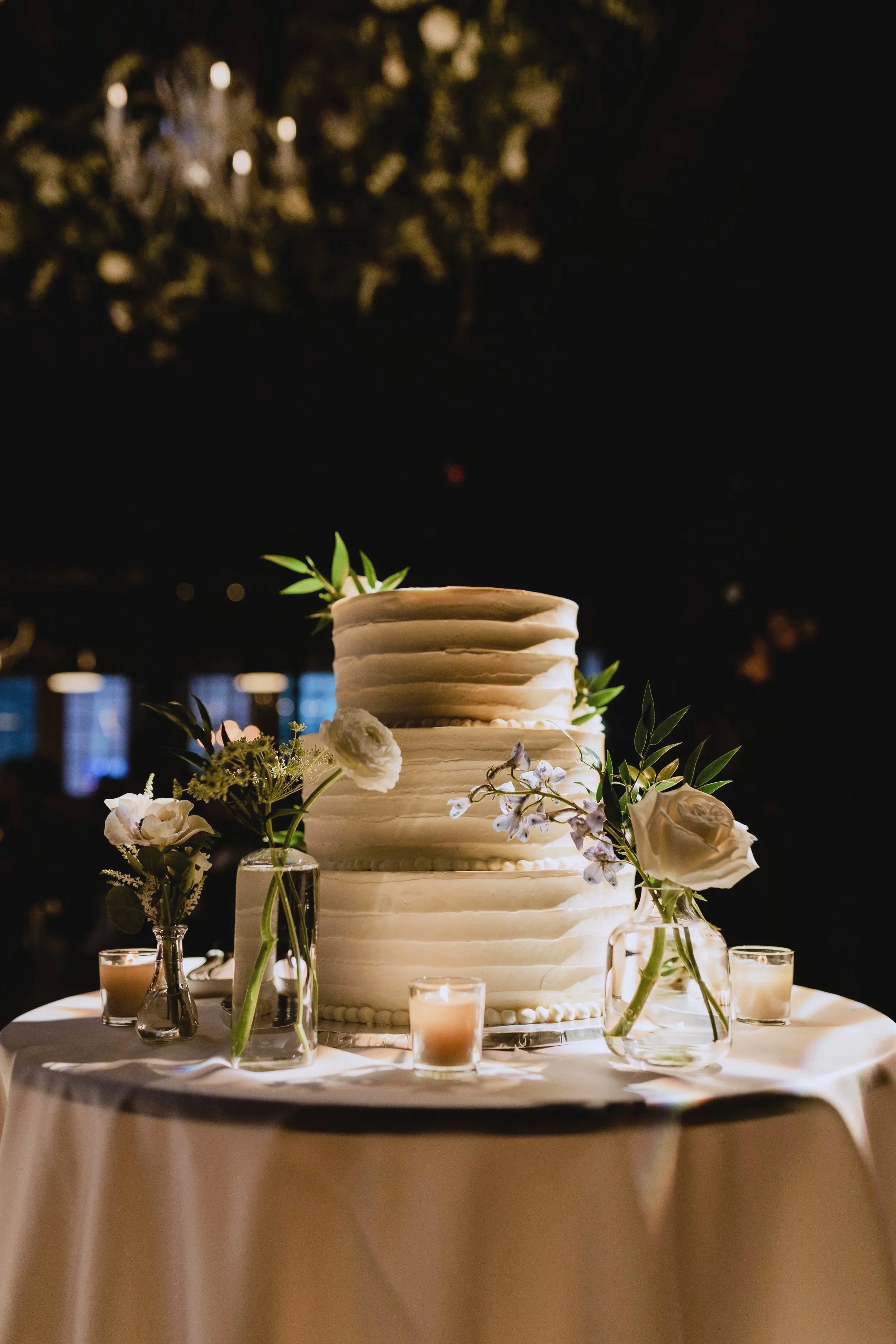 A three-tier white wedding cake with floral decorations on a table surrounded by candles and vases with flowers. Wildman BT Wedding Venue Chicago 