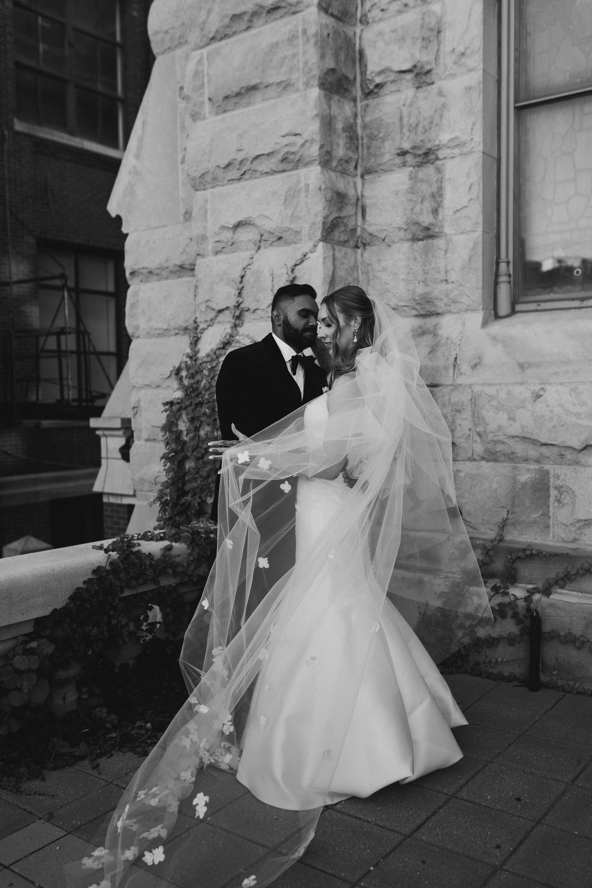 Bride and groom embracing near stone building wall, with bride's veil flowing.