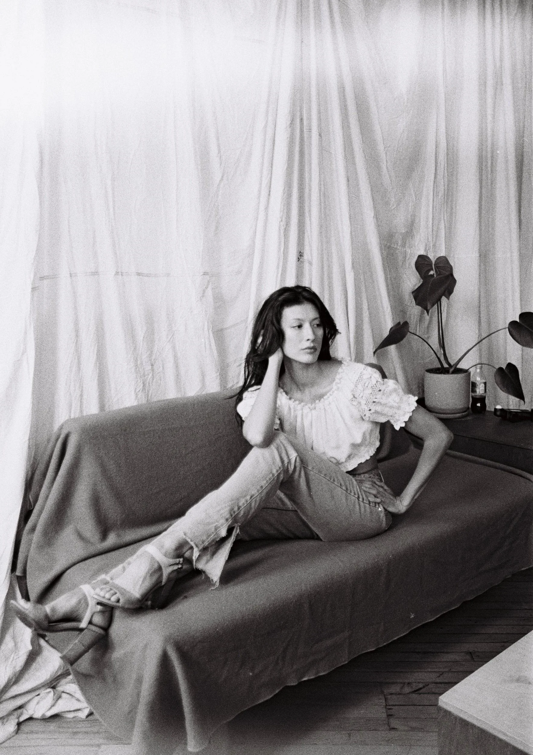 A woman sitting on a couch in a room with a curtain backdrop and a table with plants and bottles.