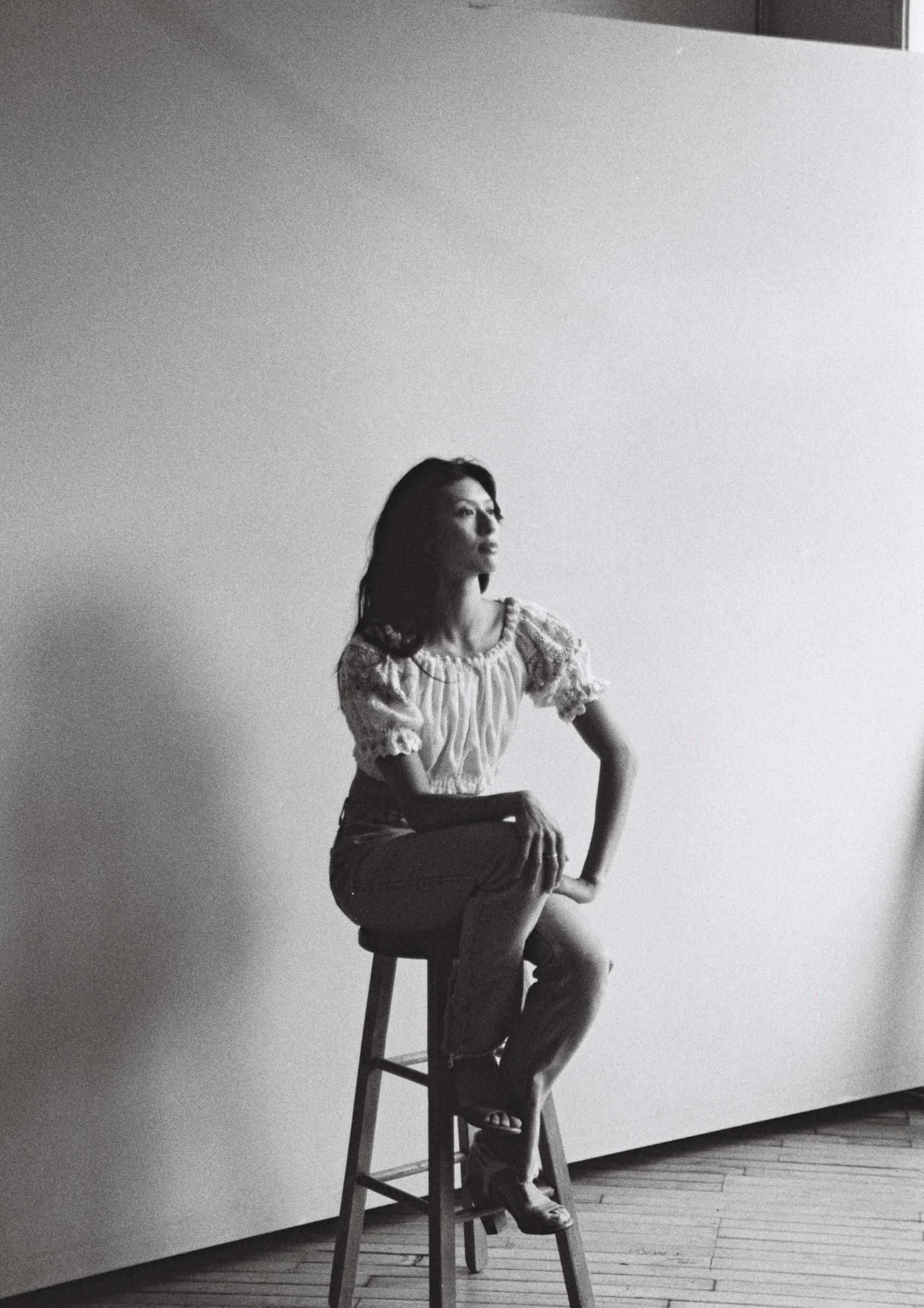 A woman with dark hair seated on a stool in front of a plain wall, appearing to be in a photo studio, in black and white.