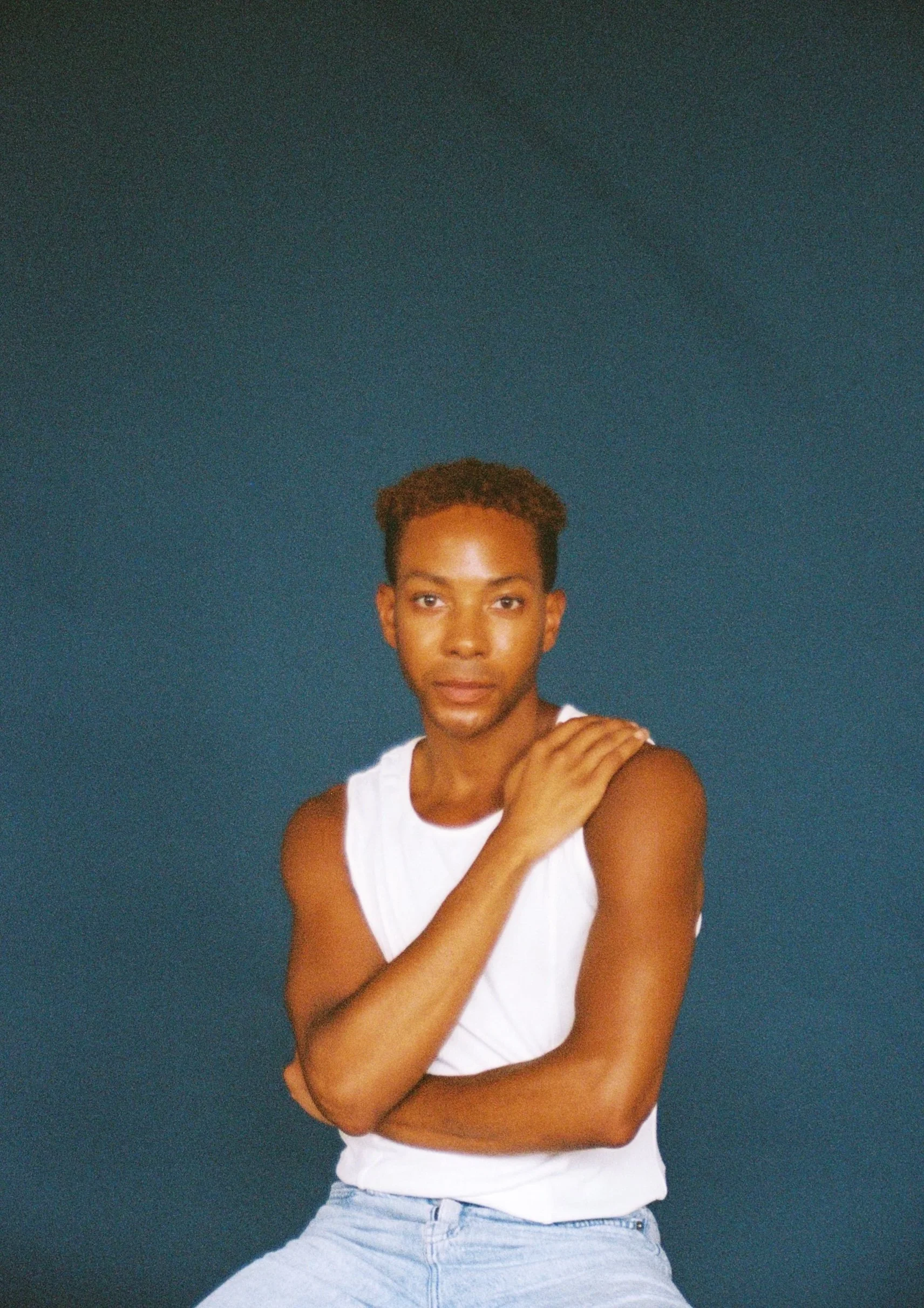 A young man with short, curly hair, wearing a white sleeveless shirt and light blue jeans, looking directly at the camera with a neutral expression, standing against a solid dark blue background.