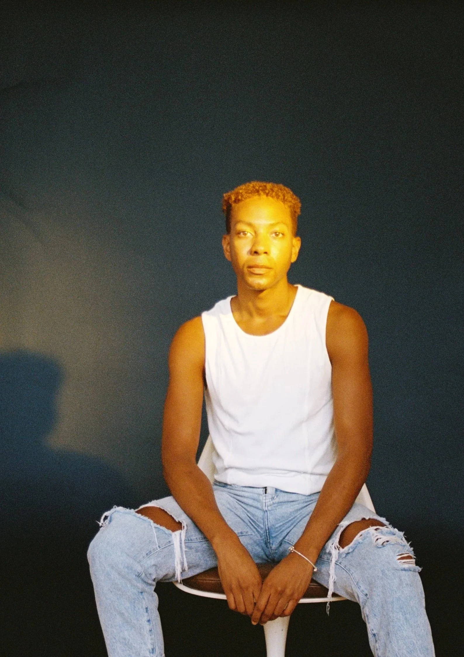 A young man with short, curly, reddish hair sitting on a chair against a dark background, wearing a white sleeveless shirt and ripped jeans.