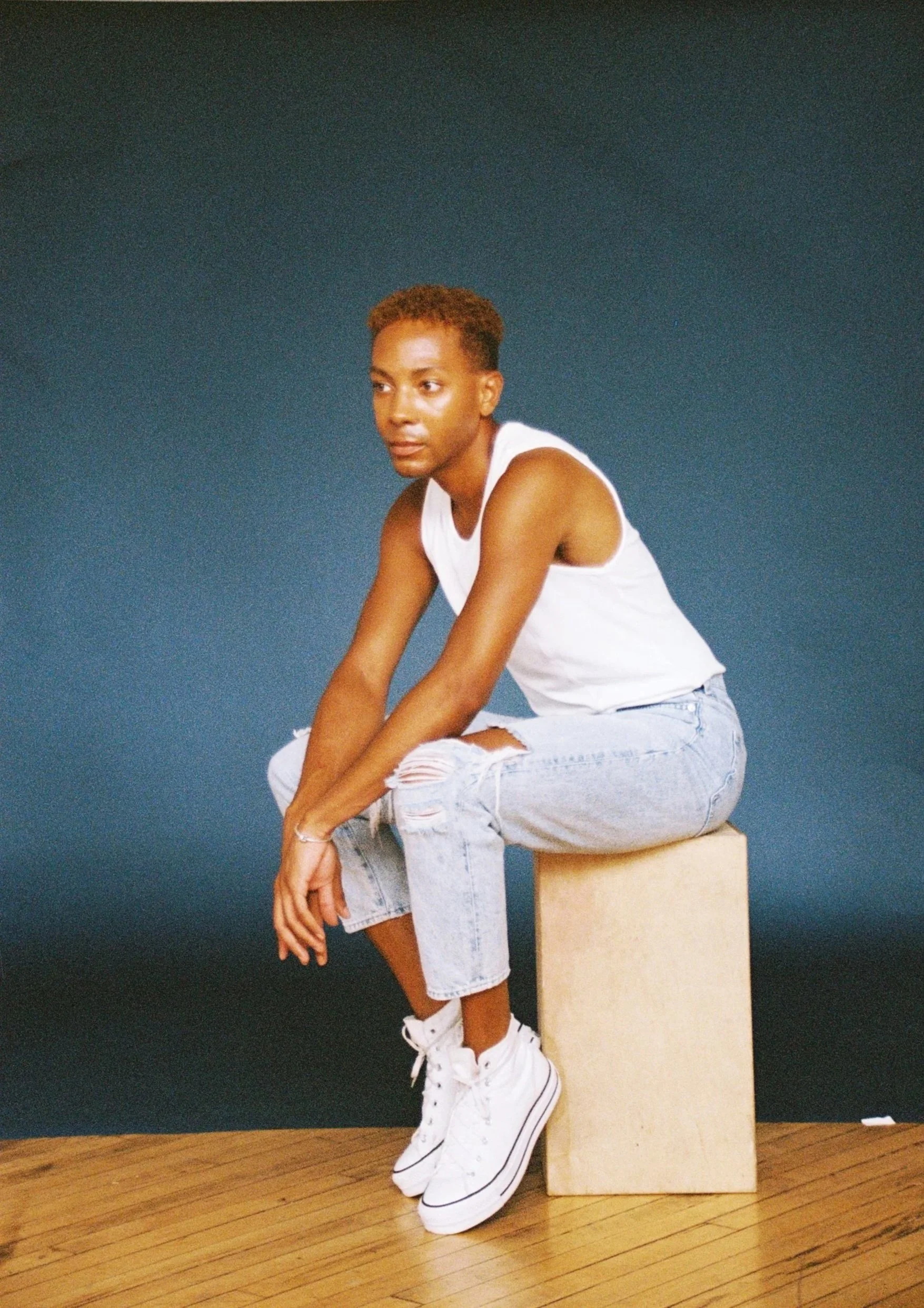 A young person with short, reddish-brown hair sitting on a wooden block, wearing a white tank top, ripped light blue jeans, and white high-top sneakers, against a dark blue background.