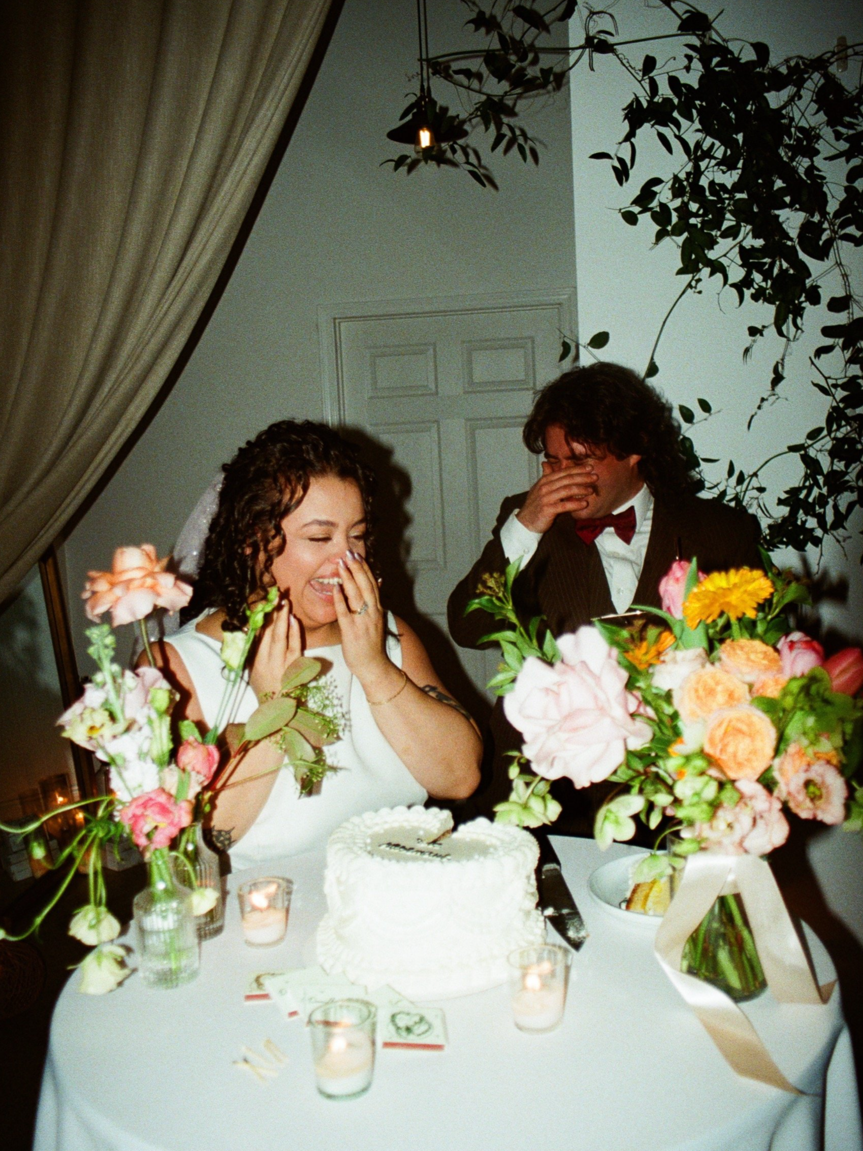 A bride and groom celebrating at their wedding reception at RLM events Chicago, standing in front of a wedding cake with floral arrangements on a table. They are laughing, with the bride covering her mouth and the groom wiping tears from his eyes.