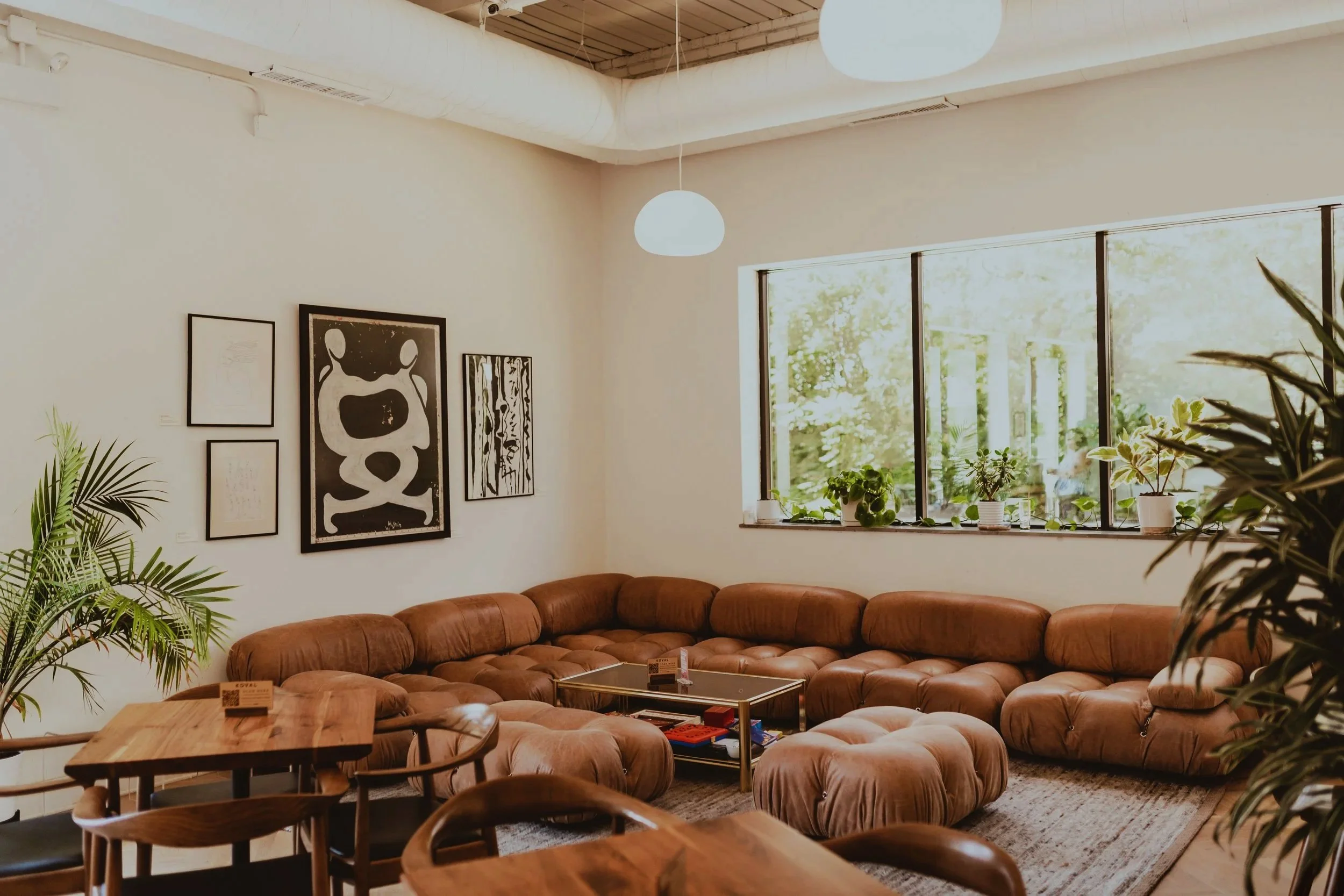 Cozy living room with a large brown sectional sofa, wooden coffee table, and potted plants near a large window with trees outside, decorated with modern art on the white wall.