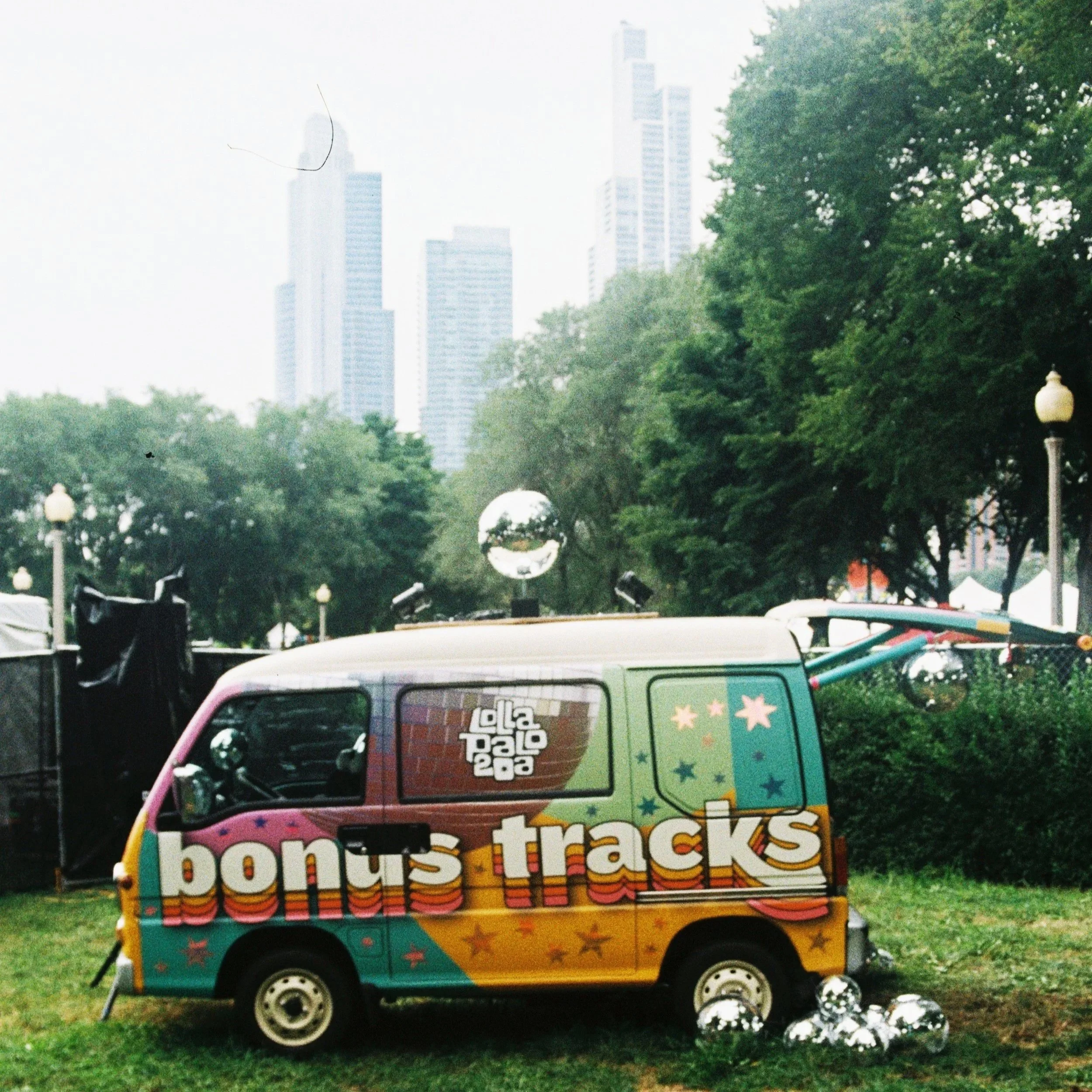Colorful van with 'bonus tracks' and star designs, parked on grass at an outdoor event with trees and city skyscrapers in the background.