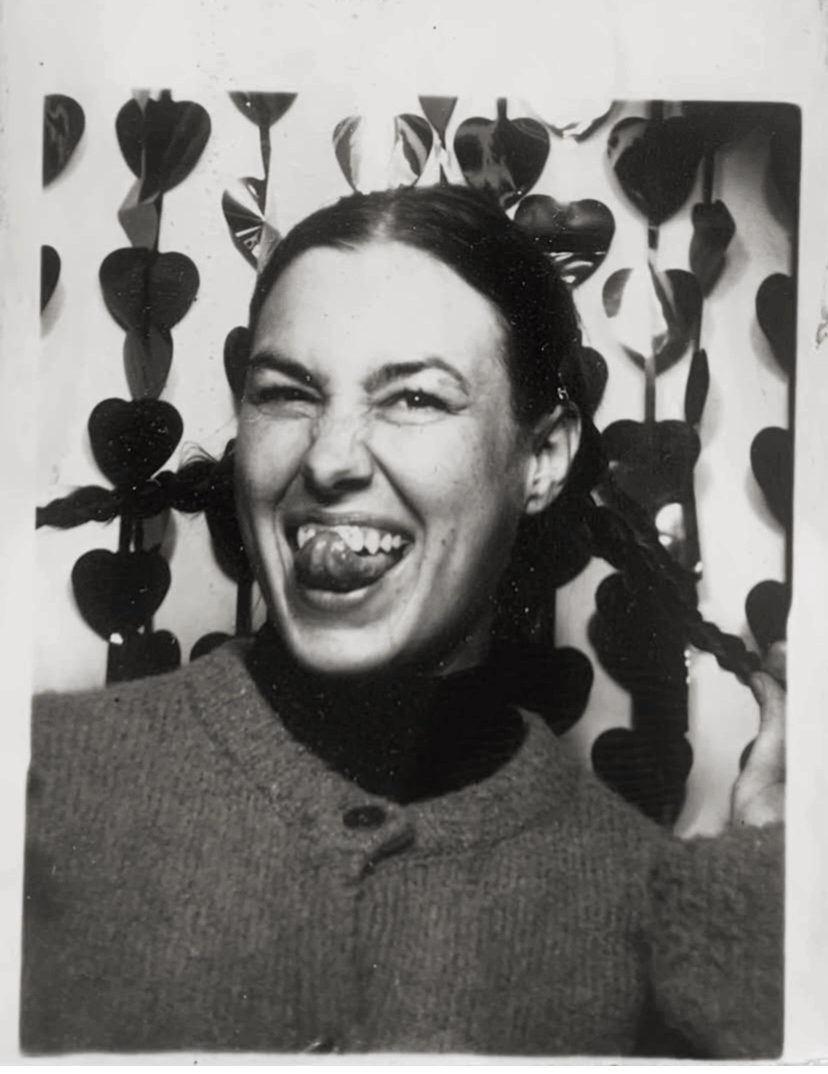 A woman with her hair in braids making a playful face with her tongue out and one eye closed, in front of a backdrop decorated with heart-shaped cutouts. Vintage Analog Photo booth Photos from The Strip Club on Damen Ave in Chicago IL