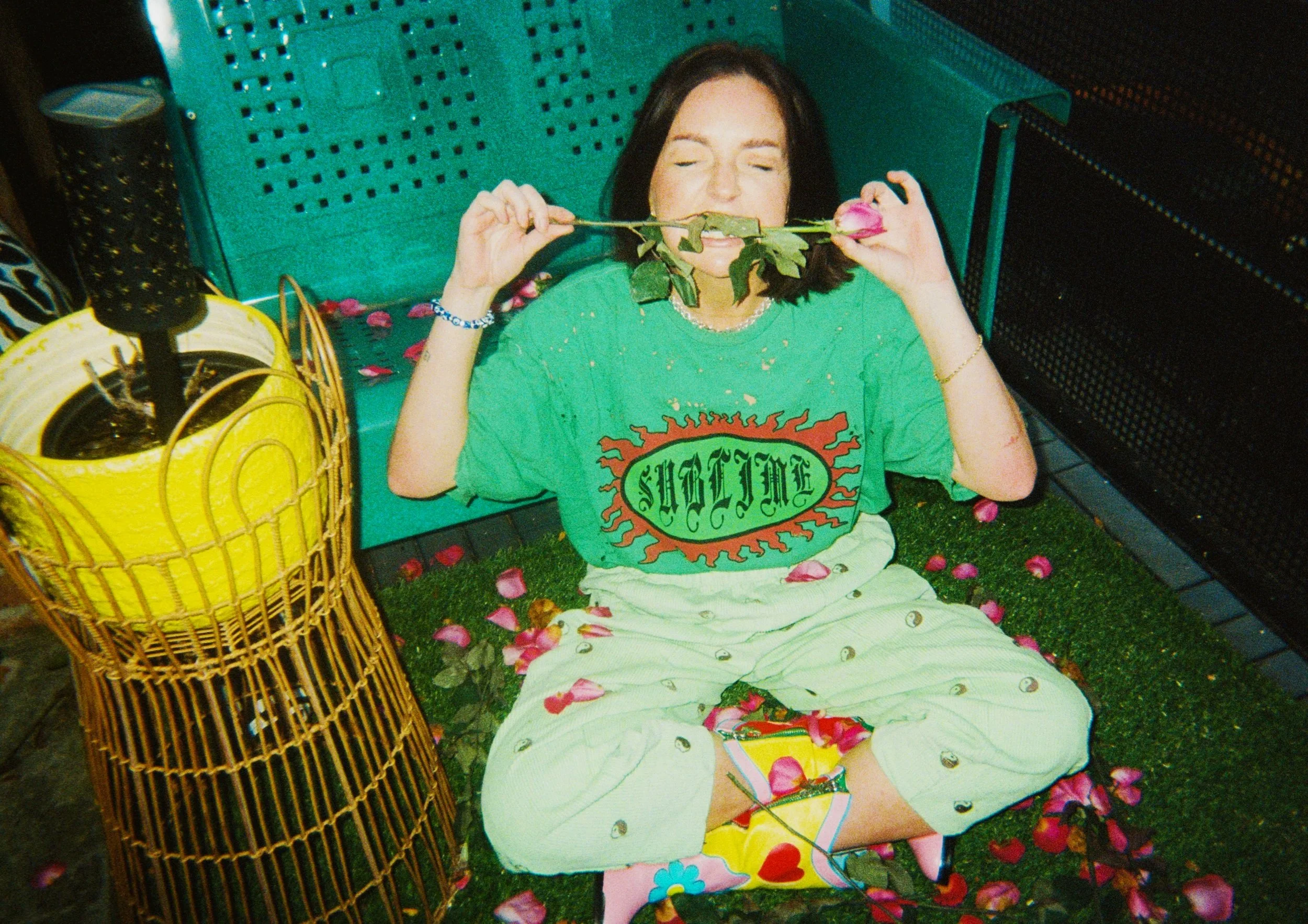 A woman sitting on the ground with her eyes closed, holding a pink rose in her mouth, surrounded by rose petals. She is wearing a green T-shirt with a graphic design and white pajama pants with a pattern. The setting includes a green bench, a yellow 