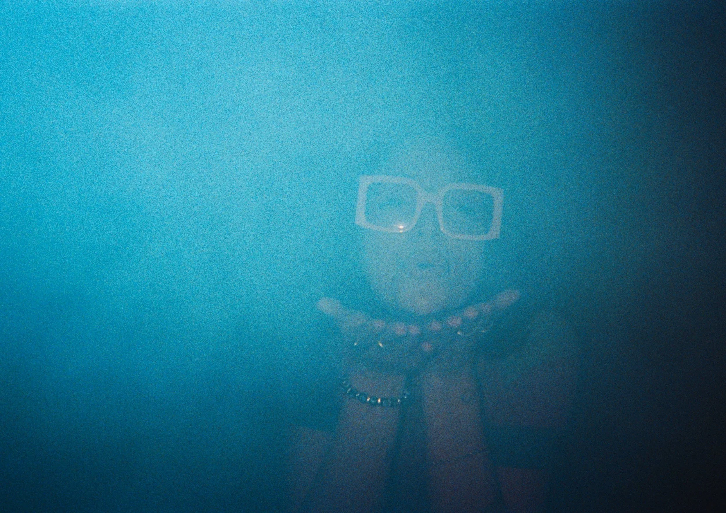 A person wearing large glasses, a jacket with a high collar, and multiple necklaces, partially obscured by dense blue fog or smoke, with only their face and upper body visible.