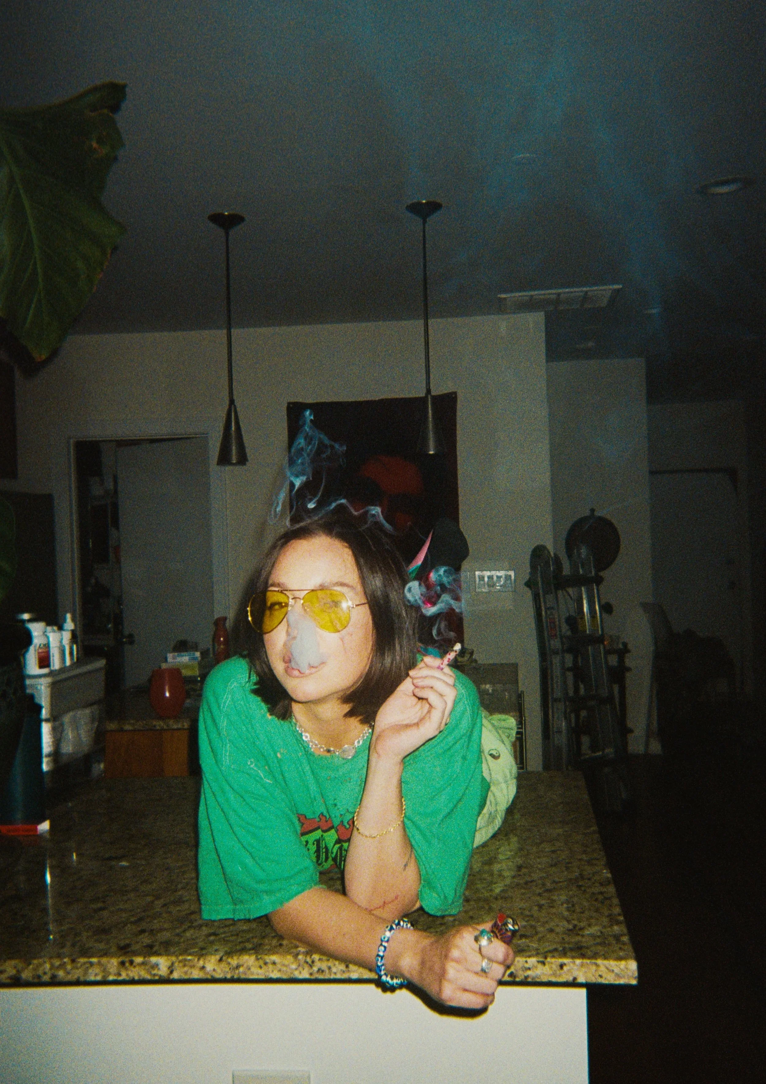 A woman with shoulder-length brown hair, yellow-tinted sunglasses, and a green shirt is smoking and leaning on a kitchen counter. Smoke is around her face, and she is wearing jewelry including rings, a bracelet, and a necklace. The kitchen has pendan