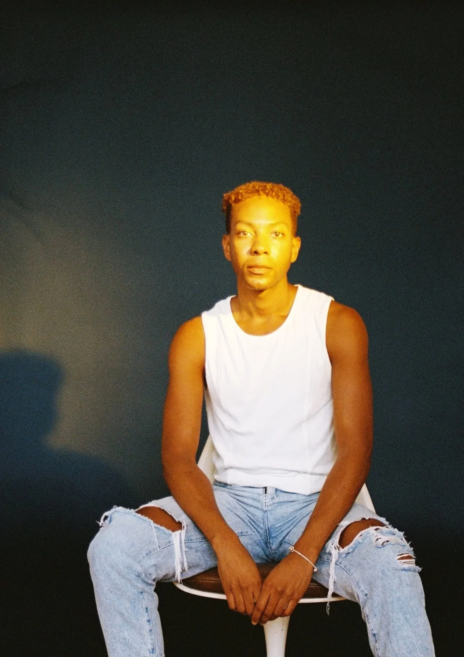 A young man with short, curly, reddish hair sitting on a chair against a dark background, wearing a white sleeveless shirt and ripped jeans.